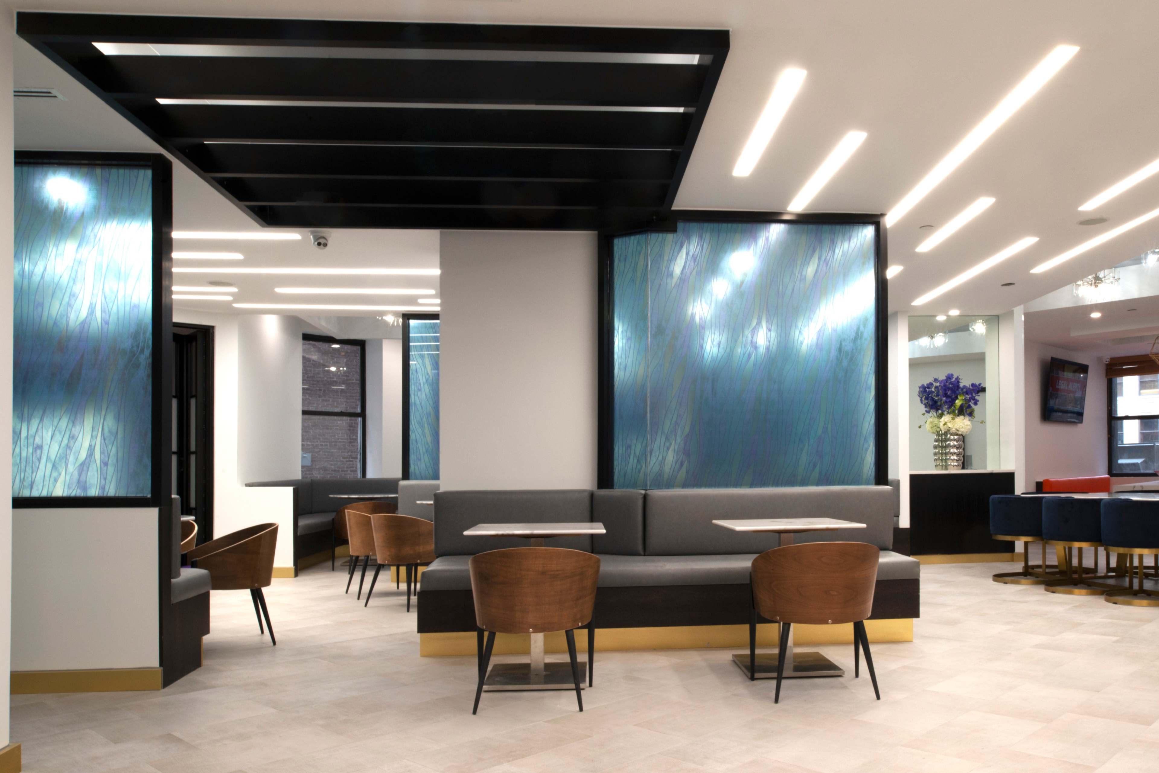 A modern café interior with seating areas featuring wooden chairs and tables, illuminated by sleek ceiling lights and decorated with blue-toned wall panels.