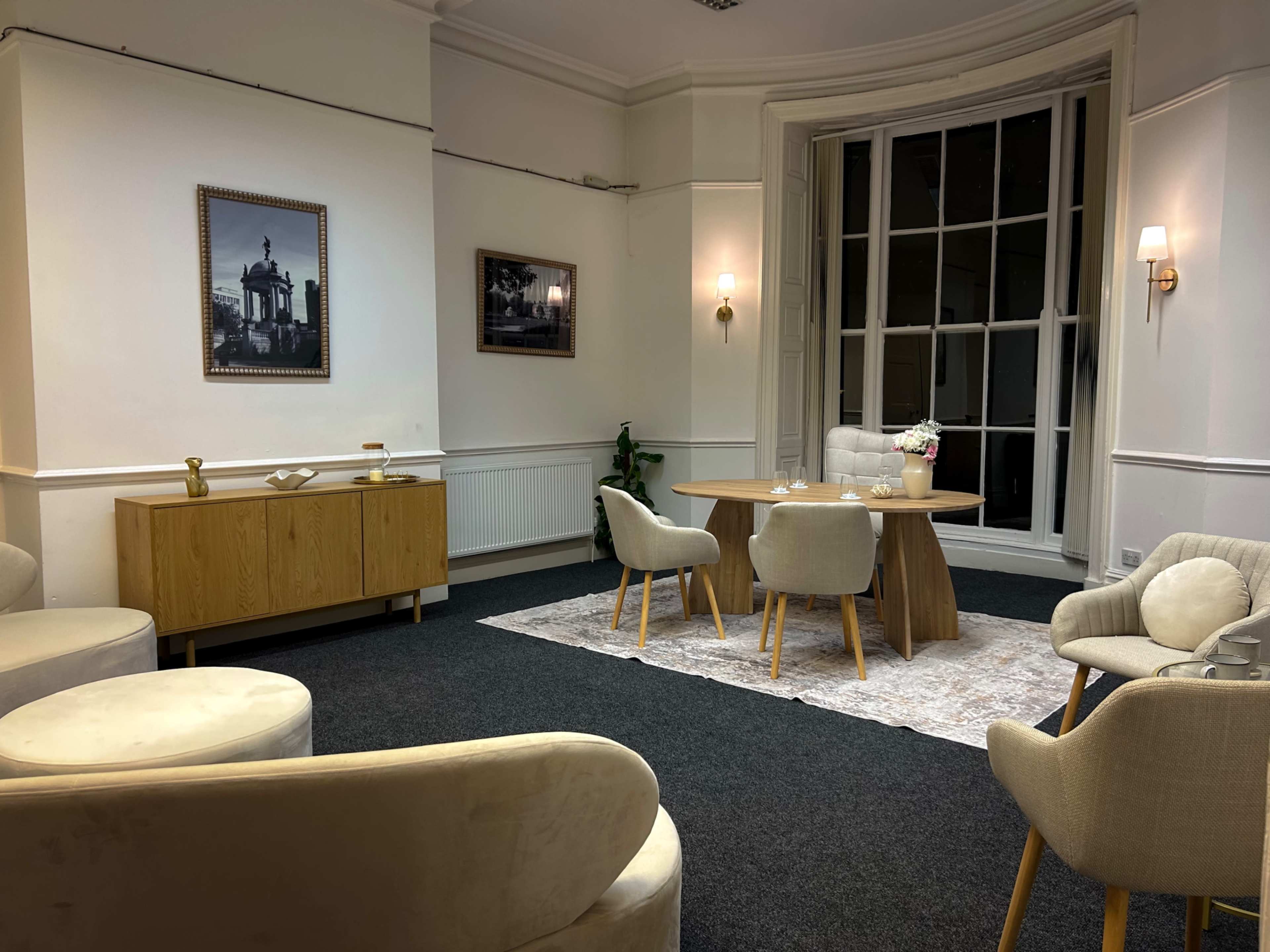 The room features a modern seating area with a round wooden table at its center, surrounded by upholstered chairs and a wooden cabinet, against a backdrop of large windows.