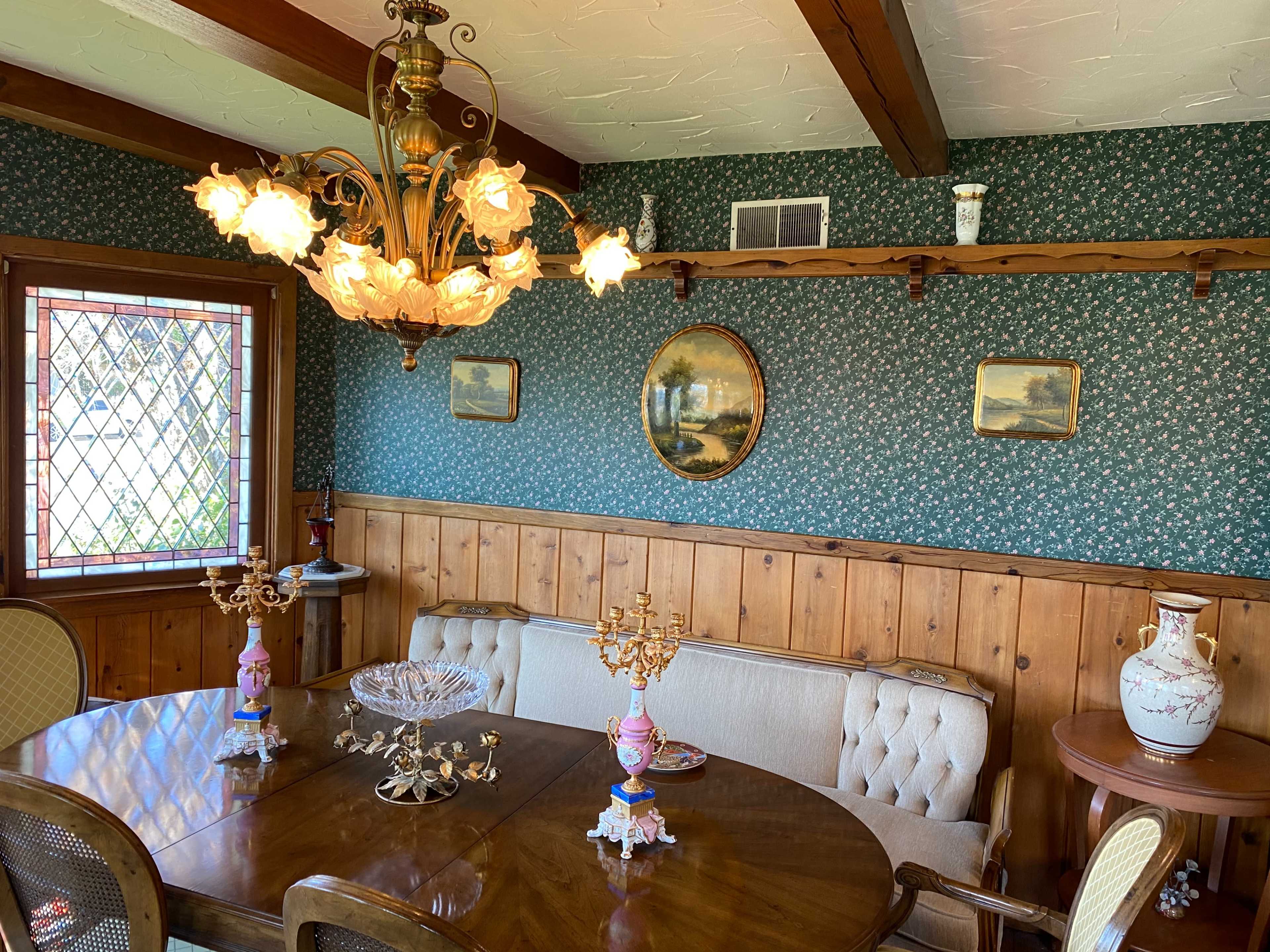 A dining room featuring a wooden table surrounded by upholstered chairs, a chandelier with floral shades hanging above, and floral wallpaper with framed paintings on the walls.