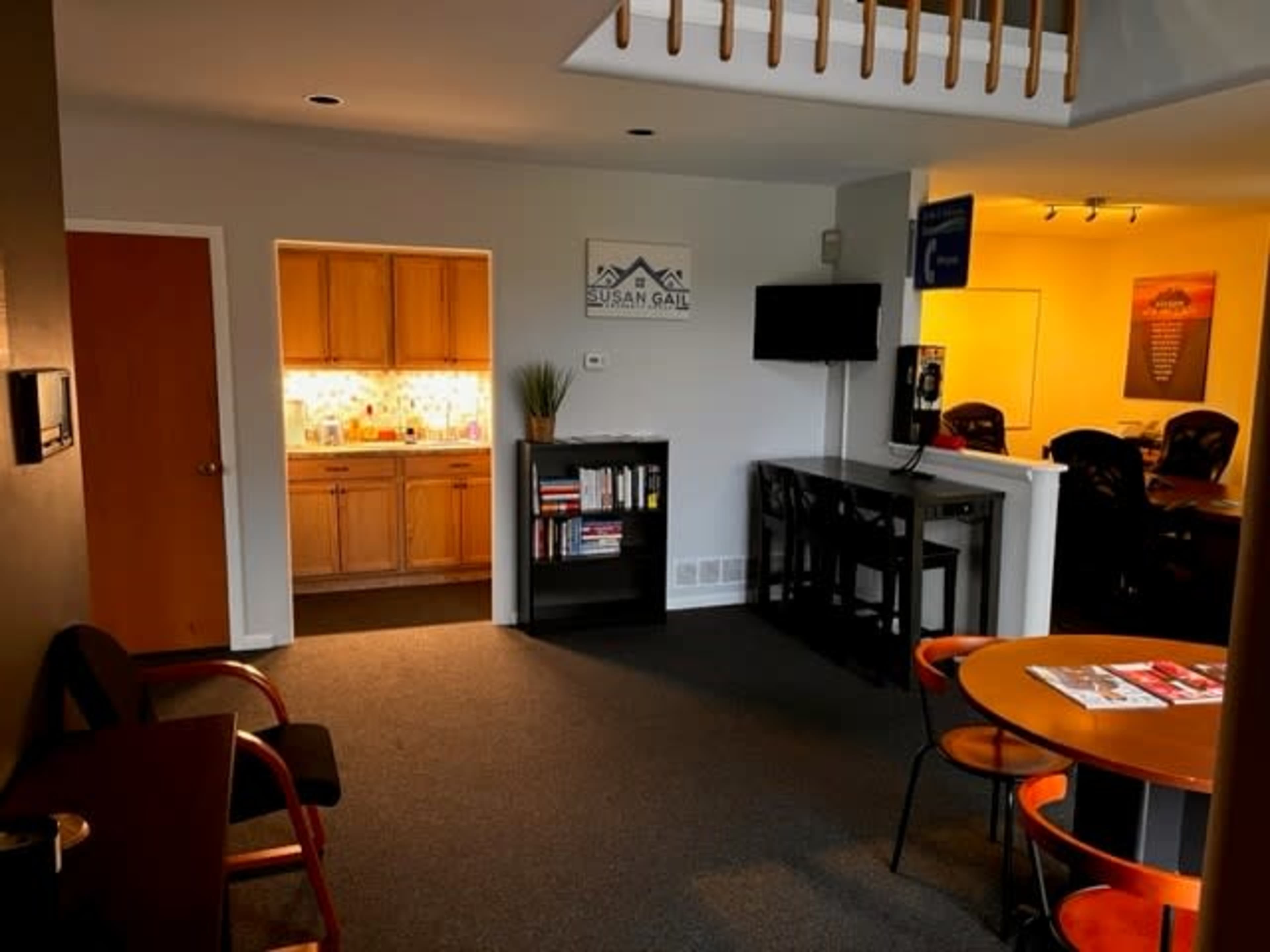 The image shows a well-lit office space with a kitchenette, bookshelves, and seating areas.