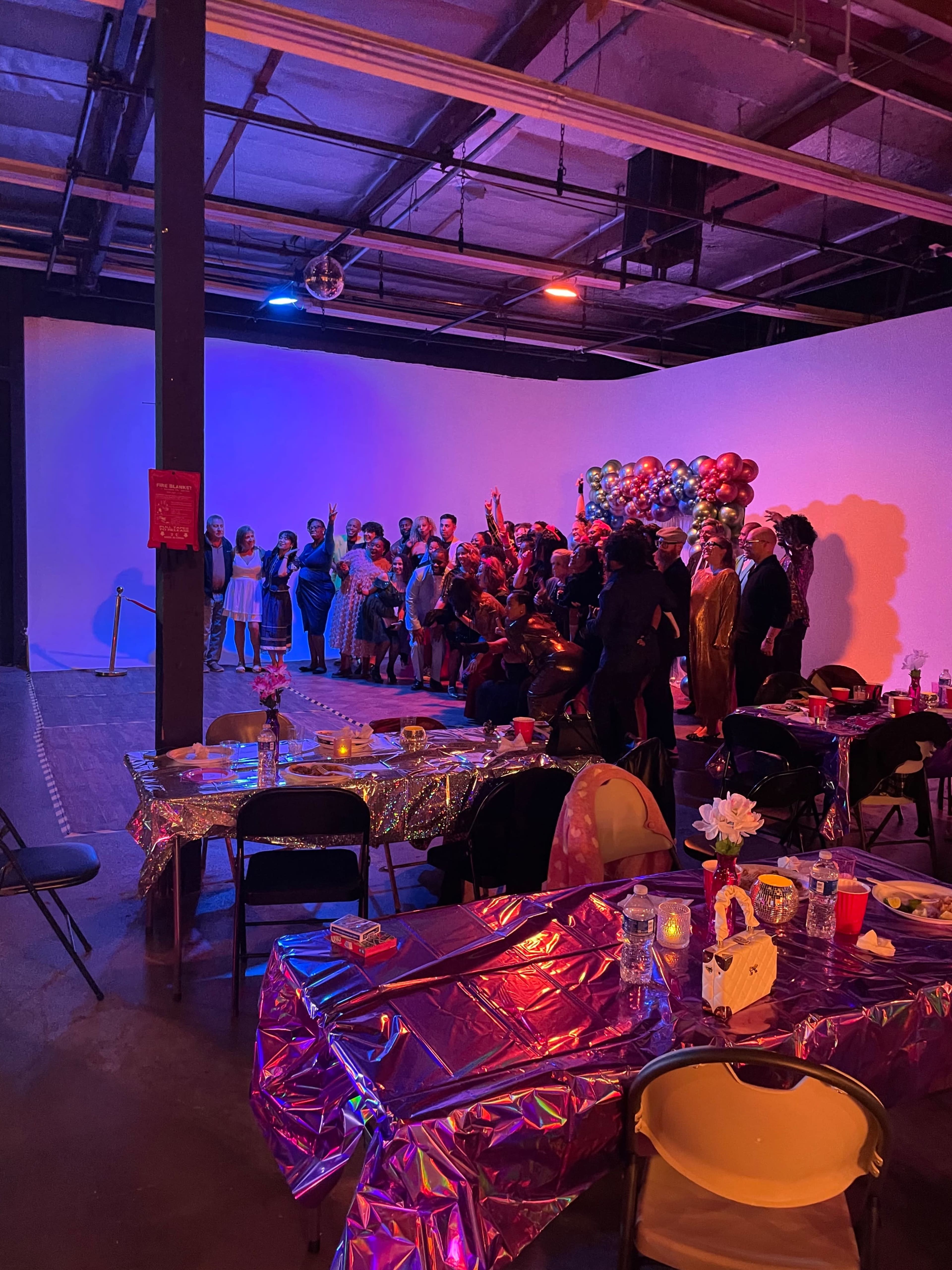 A large group of people gathers for a celebration in a dimly lit room decorated with balloons and festive tables.