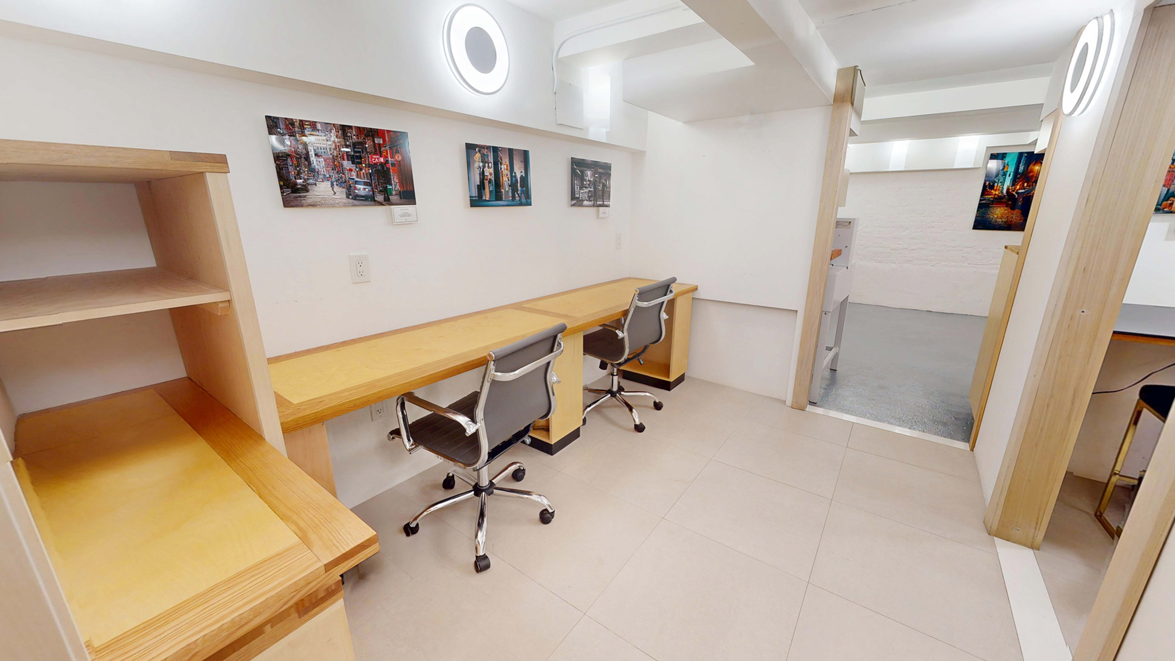 The image shows a minimalist office space with two desks and chairs positioned side by side against a white wall, featuring artwork above them.