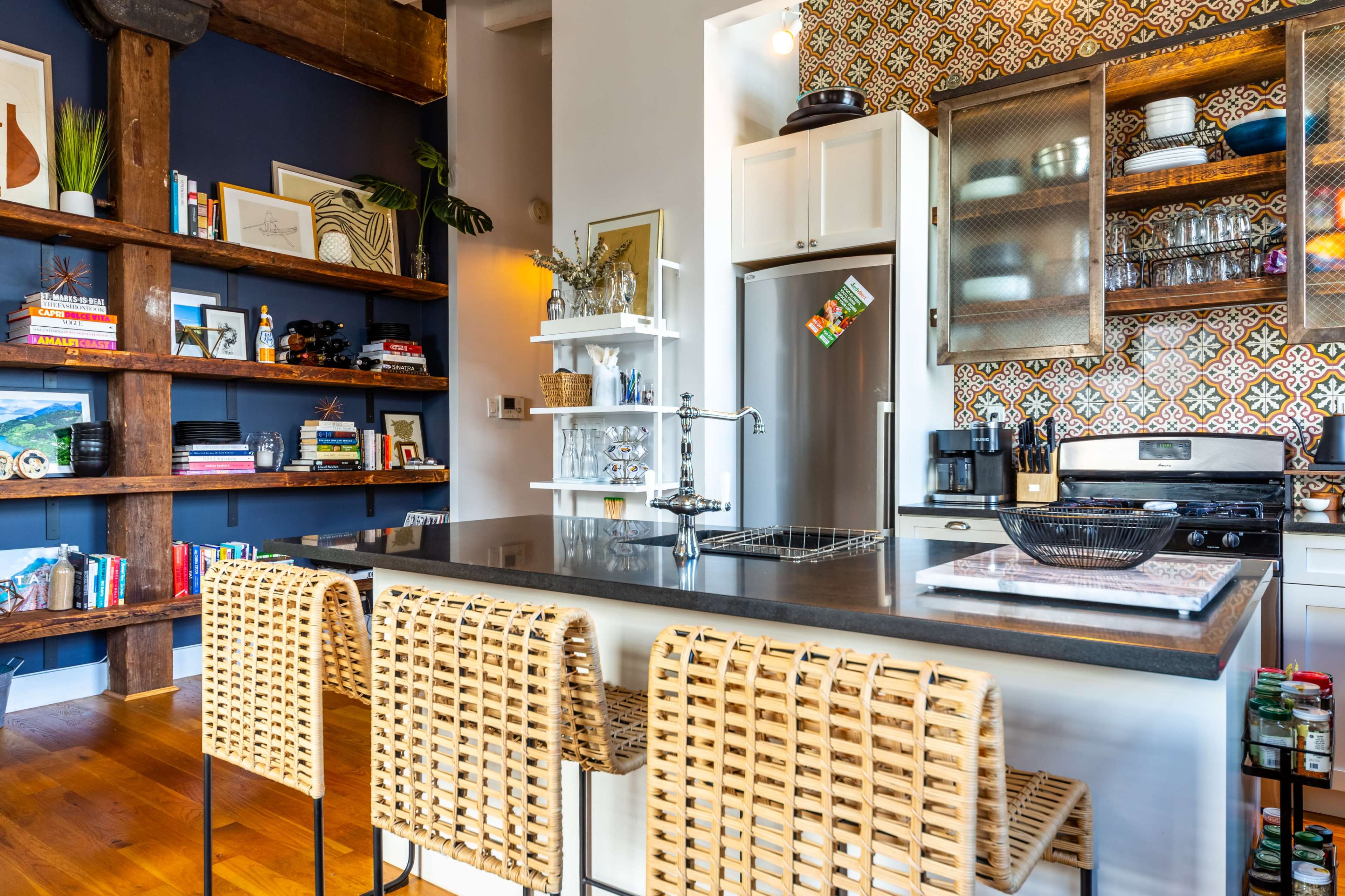 A modern kitchen with a central island, featuring wicker bar stools, open shelves filled with books and decor, and patterned tiles on the wall behind the stove.