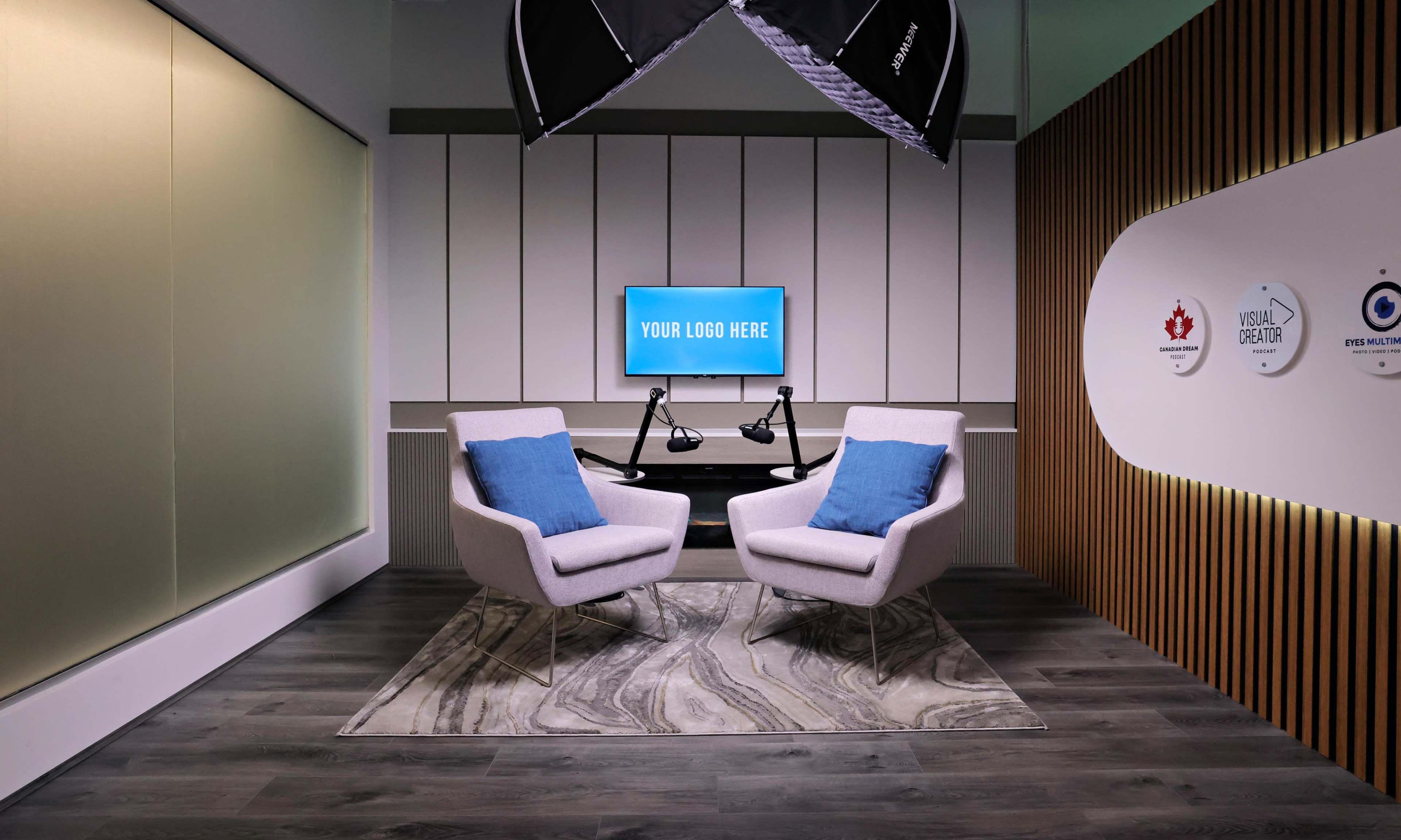 A modern studio setup features two light gray chairs with blue pillows facing a large screen displaying "YOUR LOGO HERE," surrounded by contemporary decor and lighting.