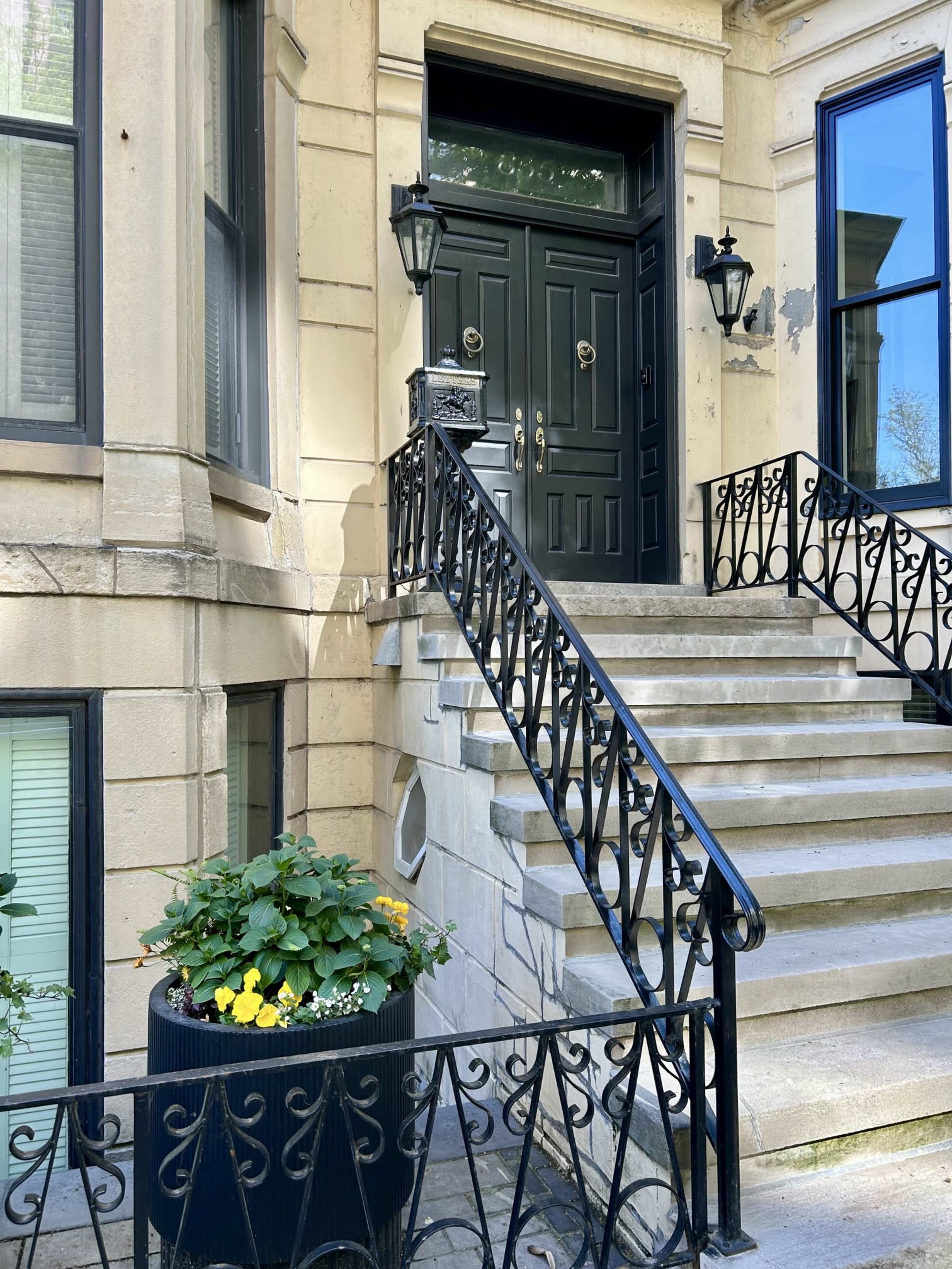 A set of stone steps with black wrought-iron railings leads up to an ornate dark front door, flanked by decorative lanterns and a planter filled with flowers.