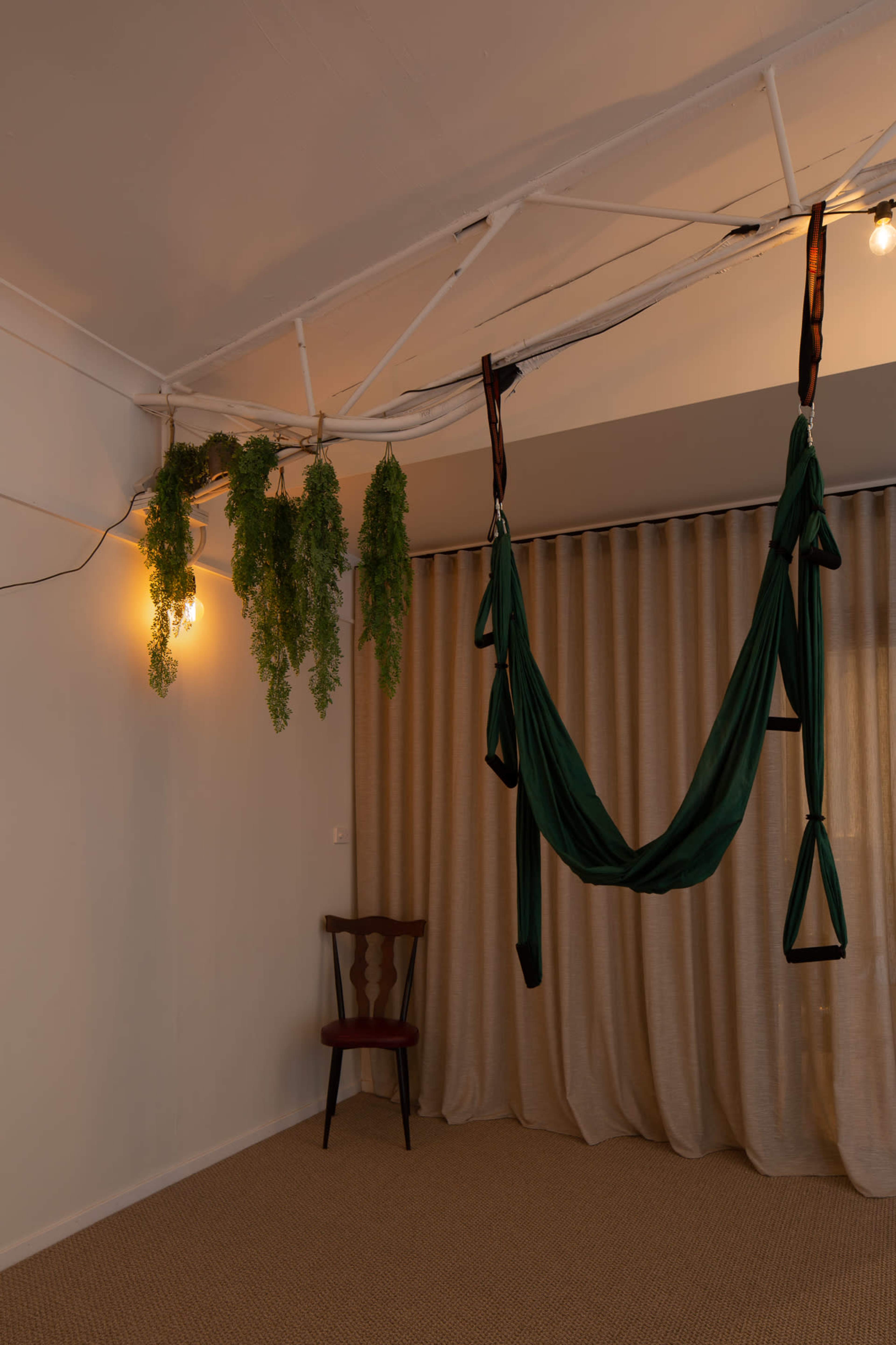 A calm indoor space featuring a green hammock suspended from the ceiling, a chair beside it, and hanging plants near a light fixture.