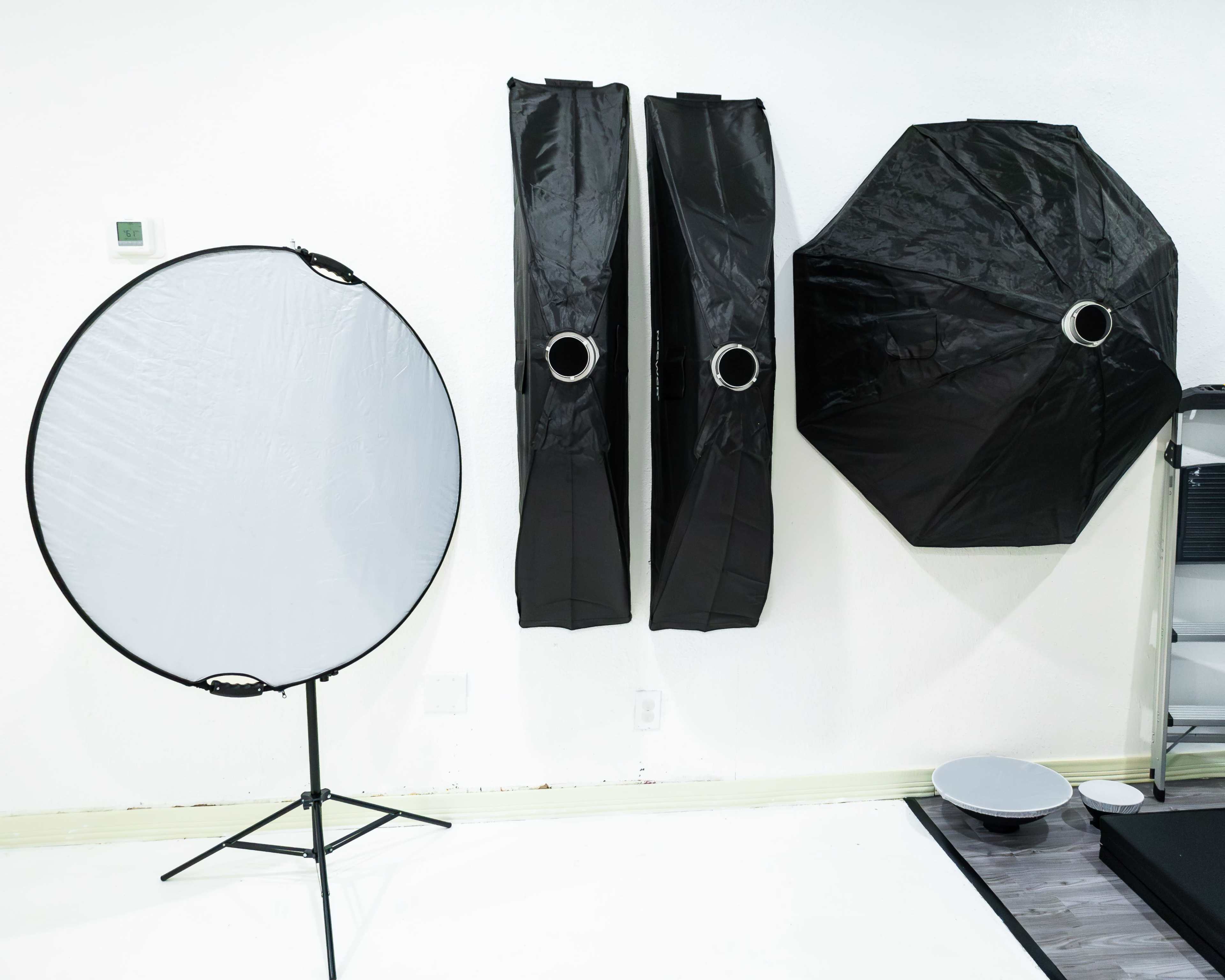 A photography setup with a round reflector on a stand next to three light diffusers arranged against a white wall.