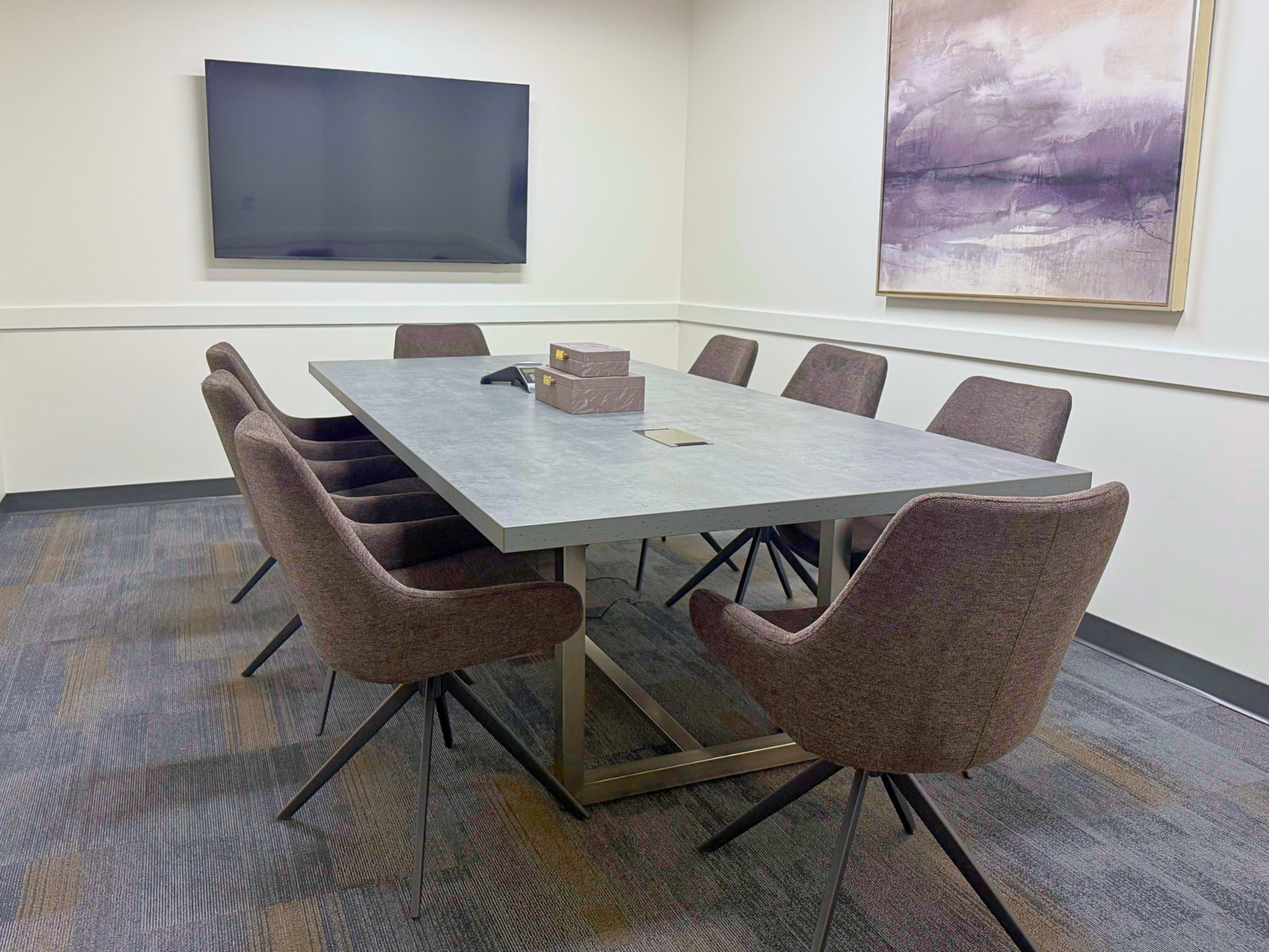 A conference room features a large gray table surrounded by eight brown chairs, with a television mounted on the wall and a piece of abstract art nearby.