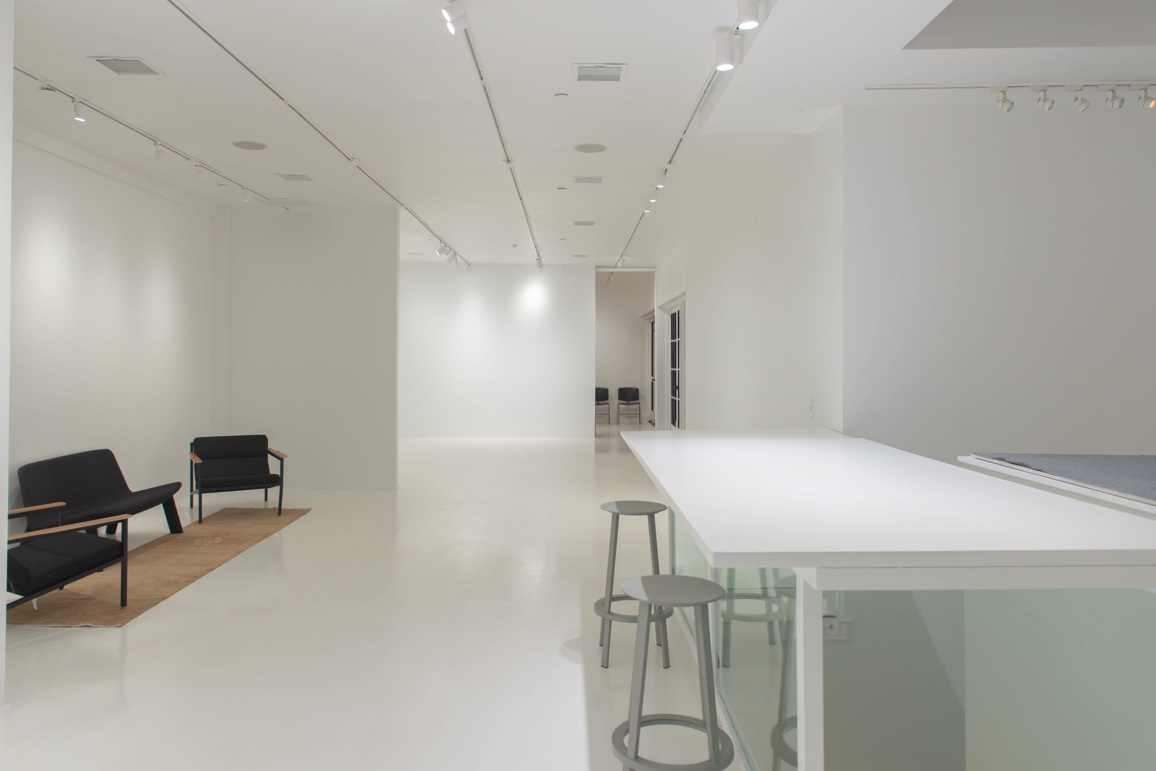 A spacious, minimalist interior with white walls and a light-colored floor, featuring a seating area with two chairs and a bar counter with stools.
