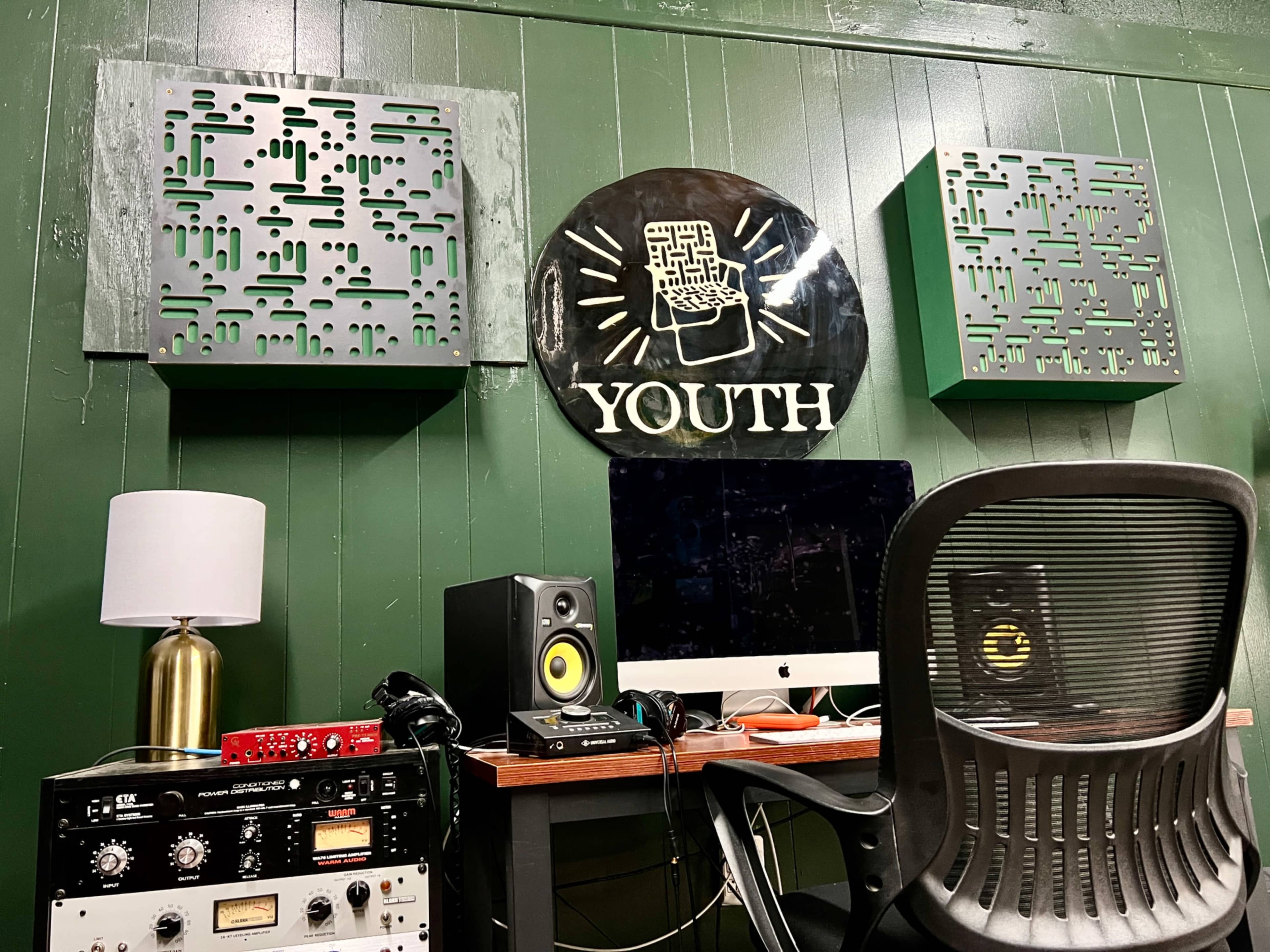 The image shows a recording studio corner with green walls, soundproof panels, a desk with a computer and speakers, and a lamp.