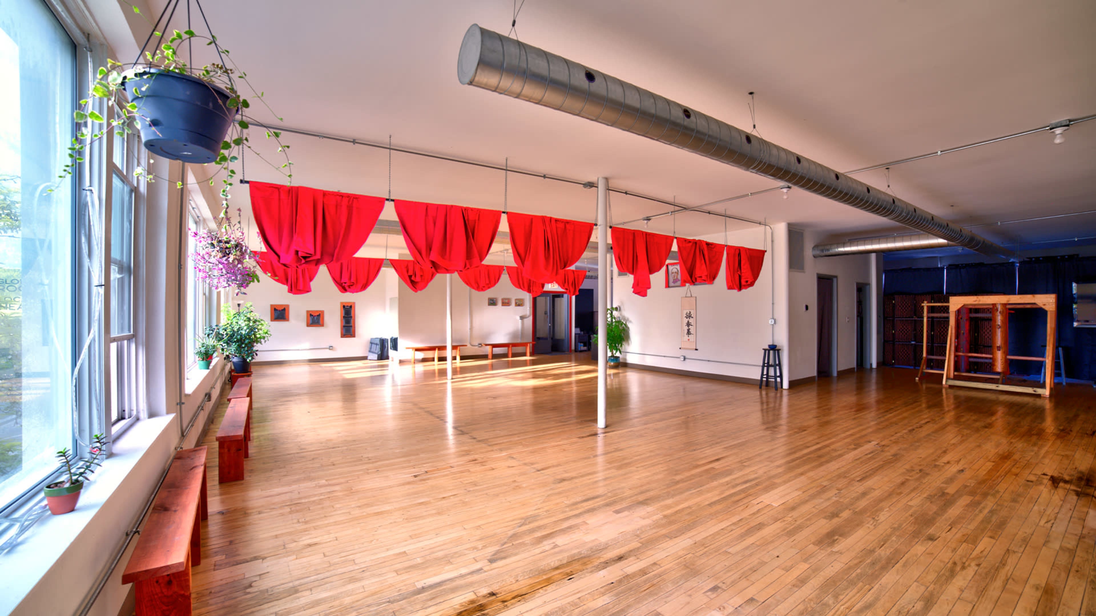 A spacious wooden-floored room with red curtains hanging from the ceiling, complemented by plants and benches along the walls.