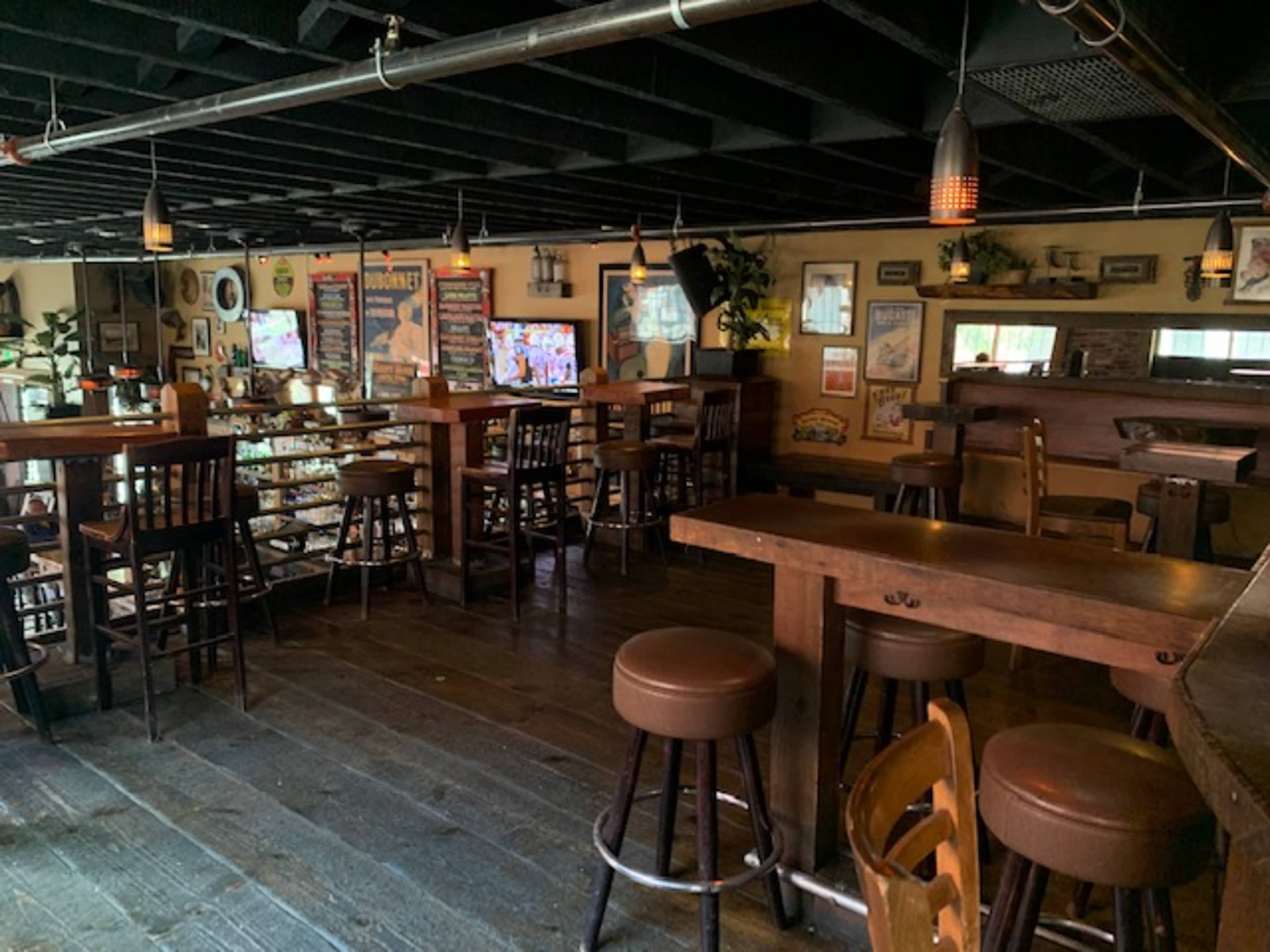 An empty bar area with wooden tables and stools, dim lighting, and televisions mounted on the walls.