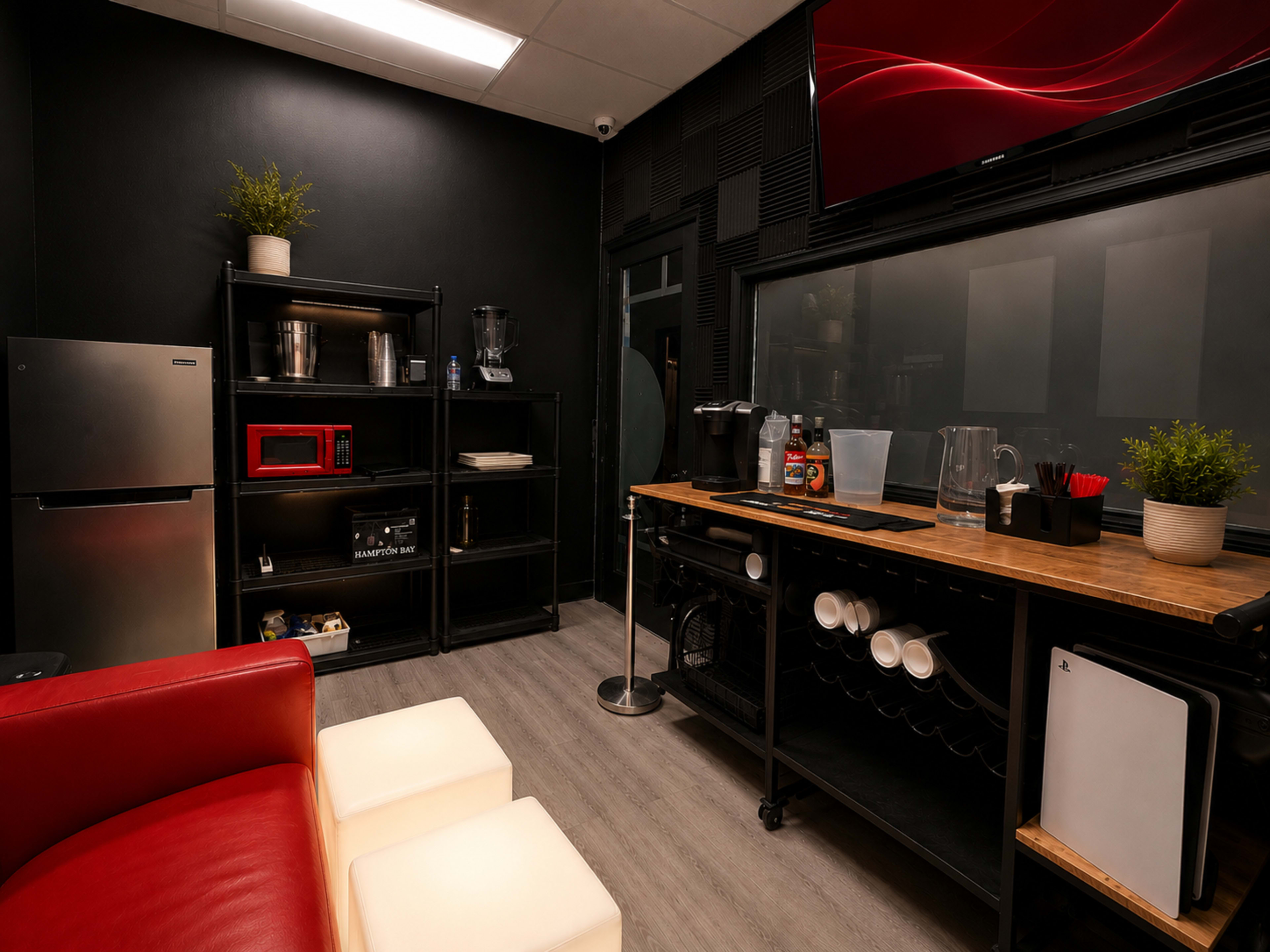 The image shows a modern break area featuring a red couch, a white table, a black shelving unit with kitchen appliances, and a countertop with various beverage supplies.