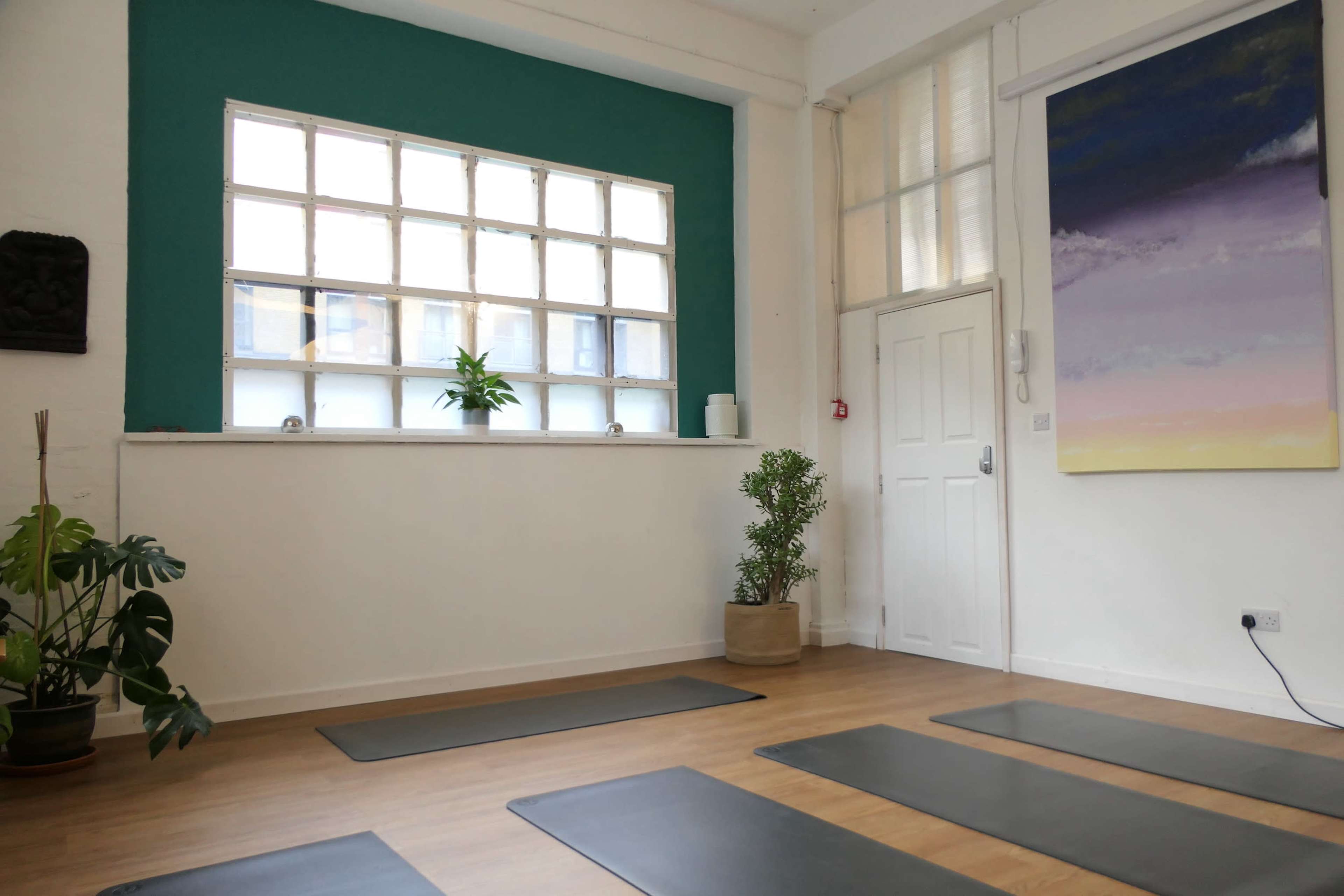 A yoga studio with several mats arranged on the wooden floor, a large window on the wall, and green plants by the window.