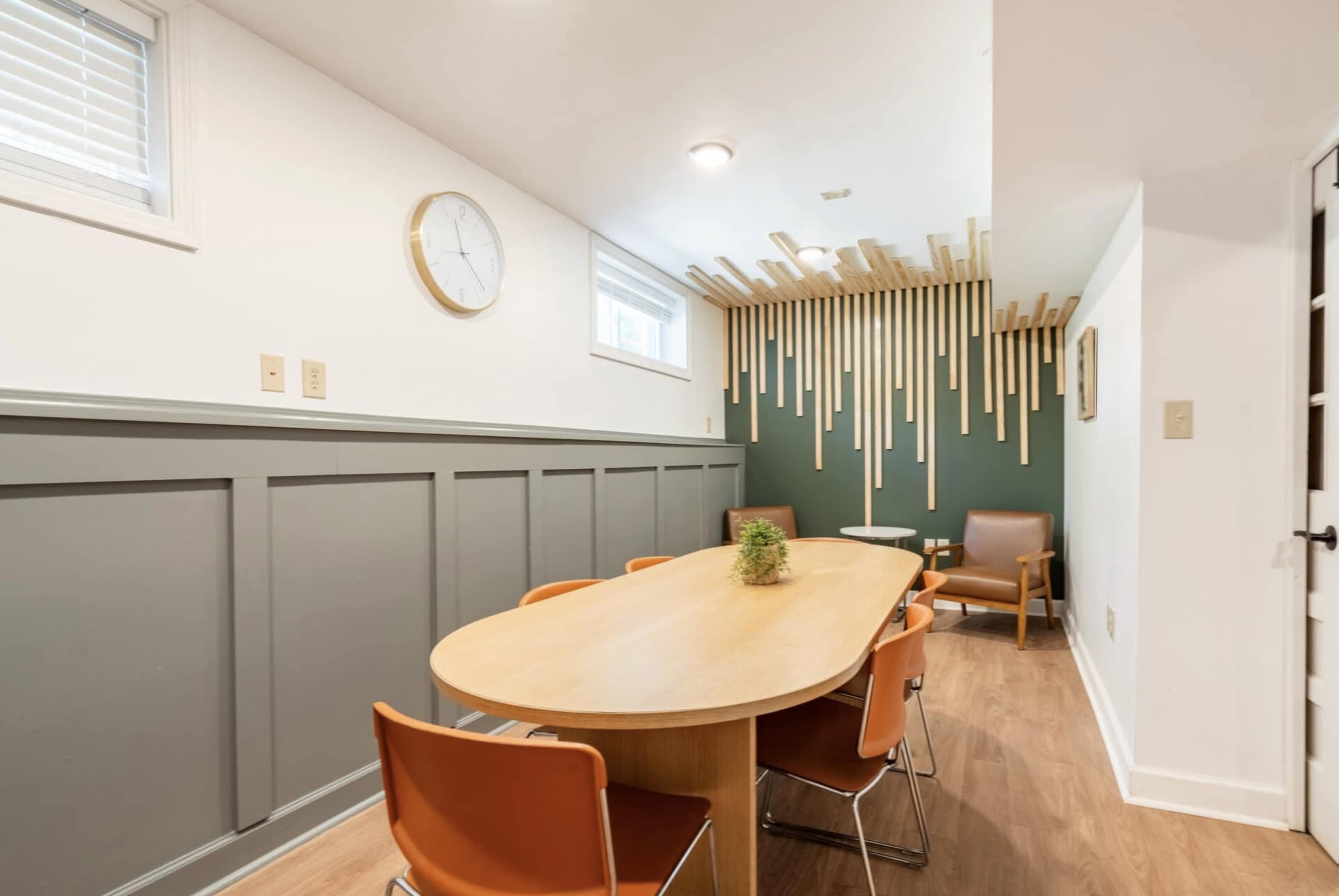 A conference room features a long wooden table surrounded by orange chairs, a wall clock, and decorative wooden slats on a green accent wall.