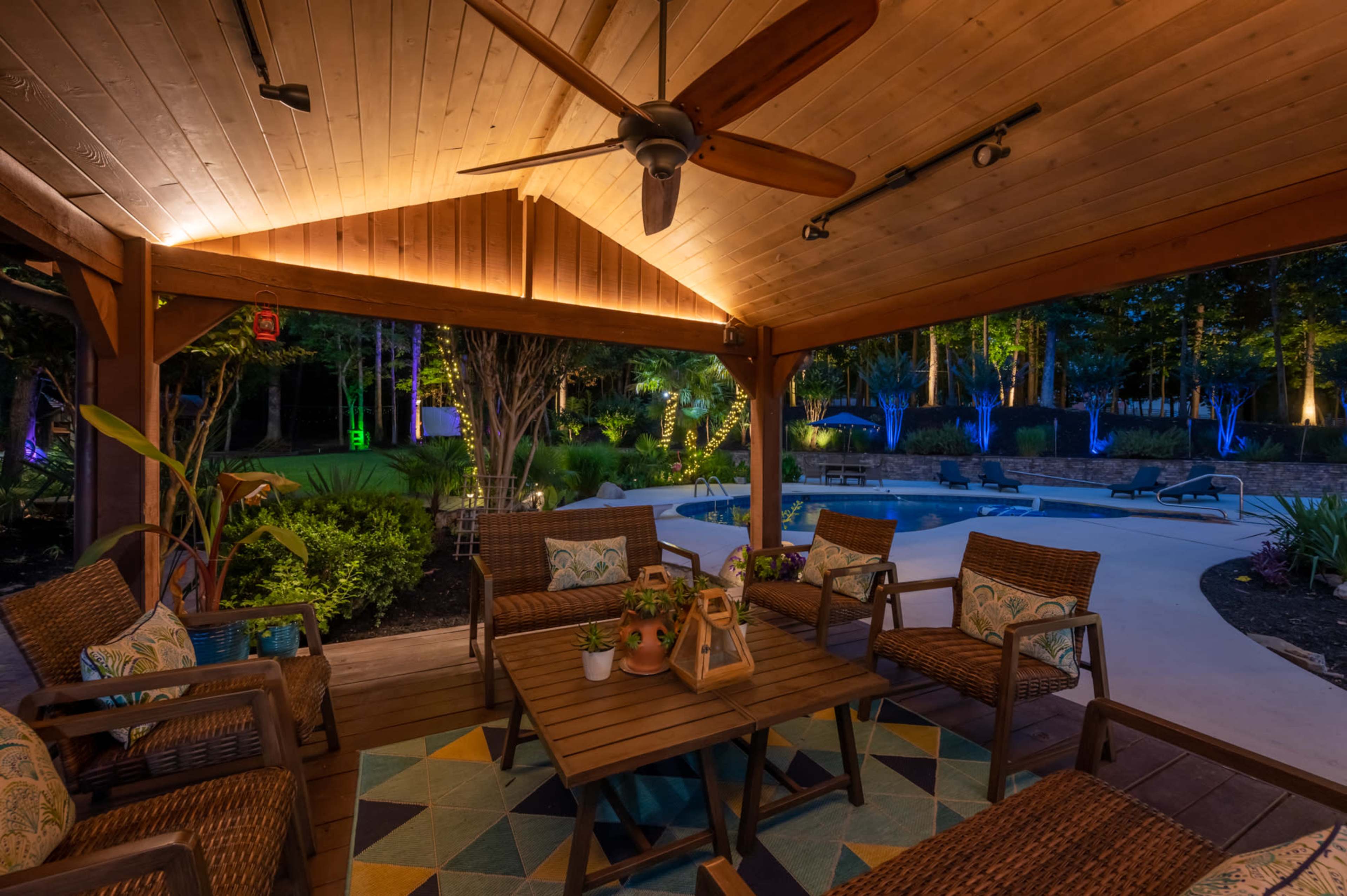 A covered outdoor seating area features wicker chairs and a wooden table, surrounded by a well-lit garden and a pool area.