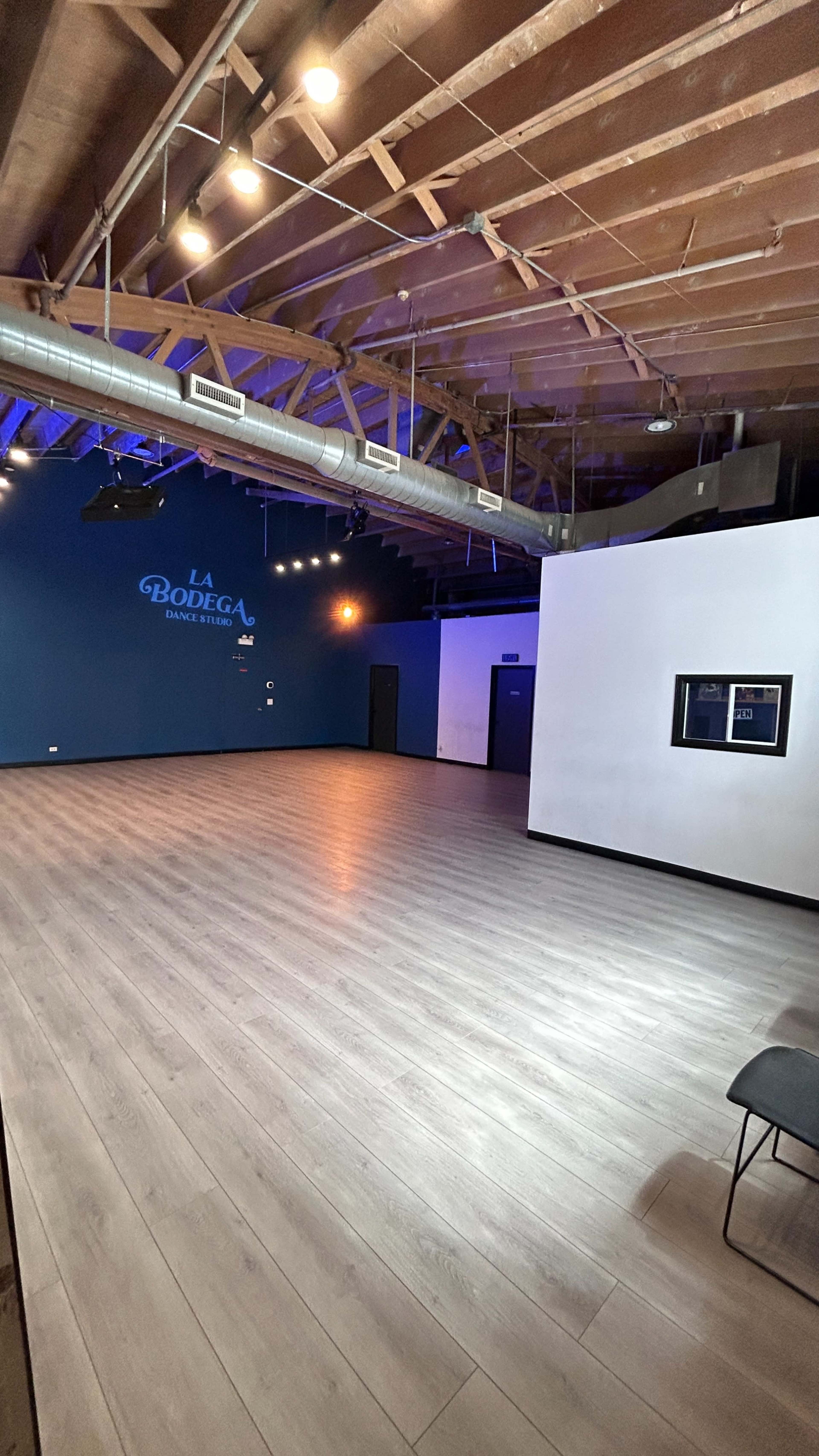 The image shows an empty, modern event space with wooden floors and dark blue and white walls, featuring the words "LA BODEGA" on one wall.