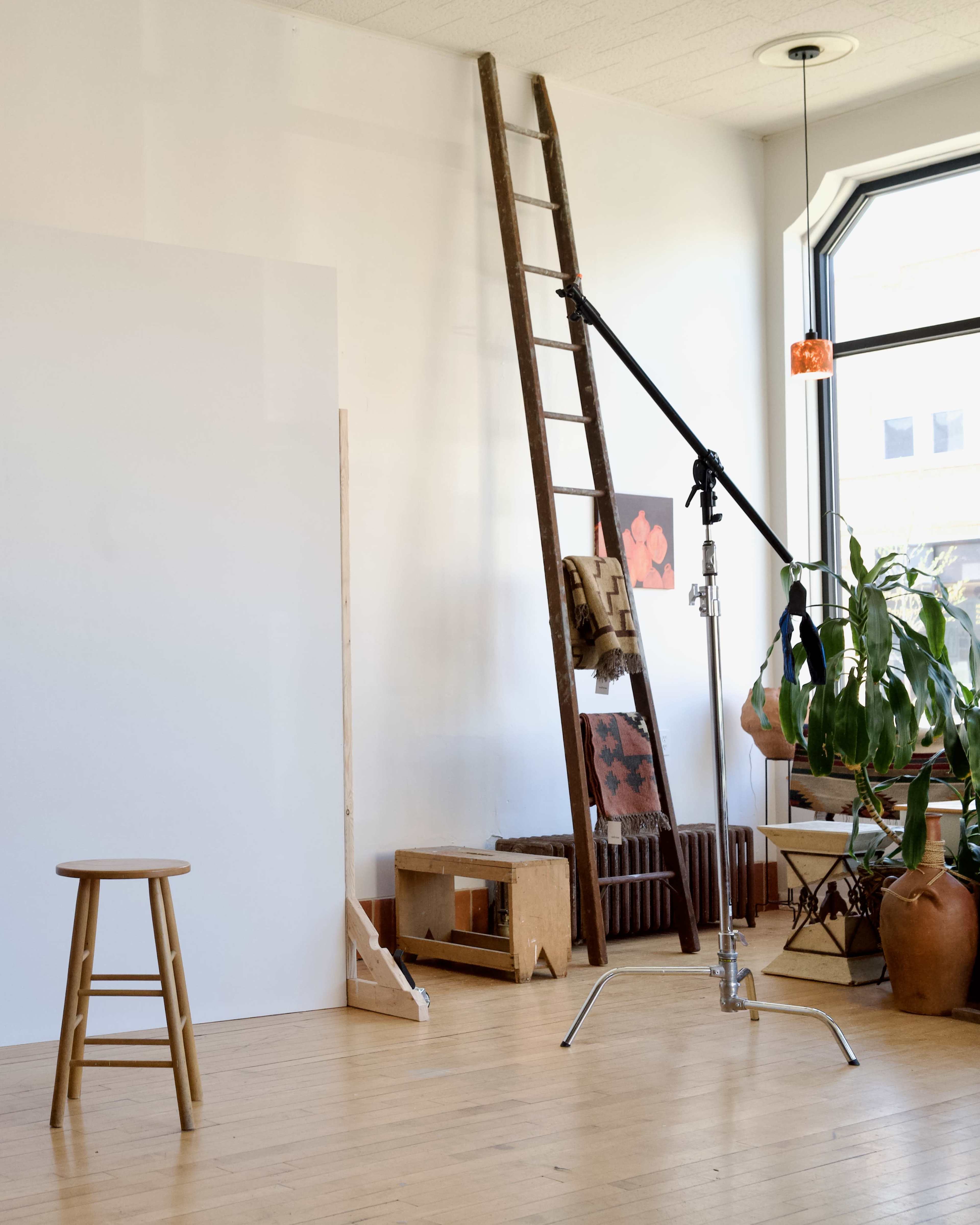 The image shows a spacious studio with a wooden ladder, a stool, a backdrop, a lighting stand, and various pieces of furniture and plants.