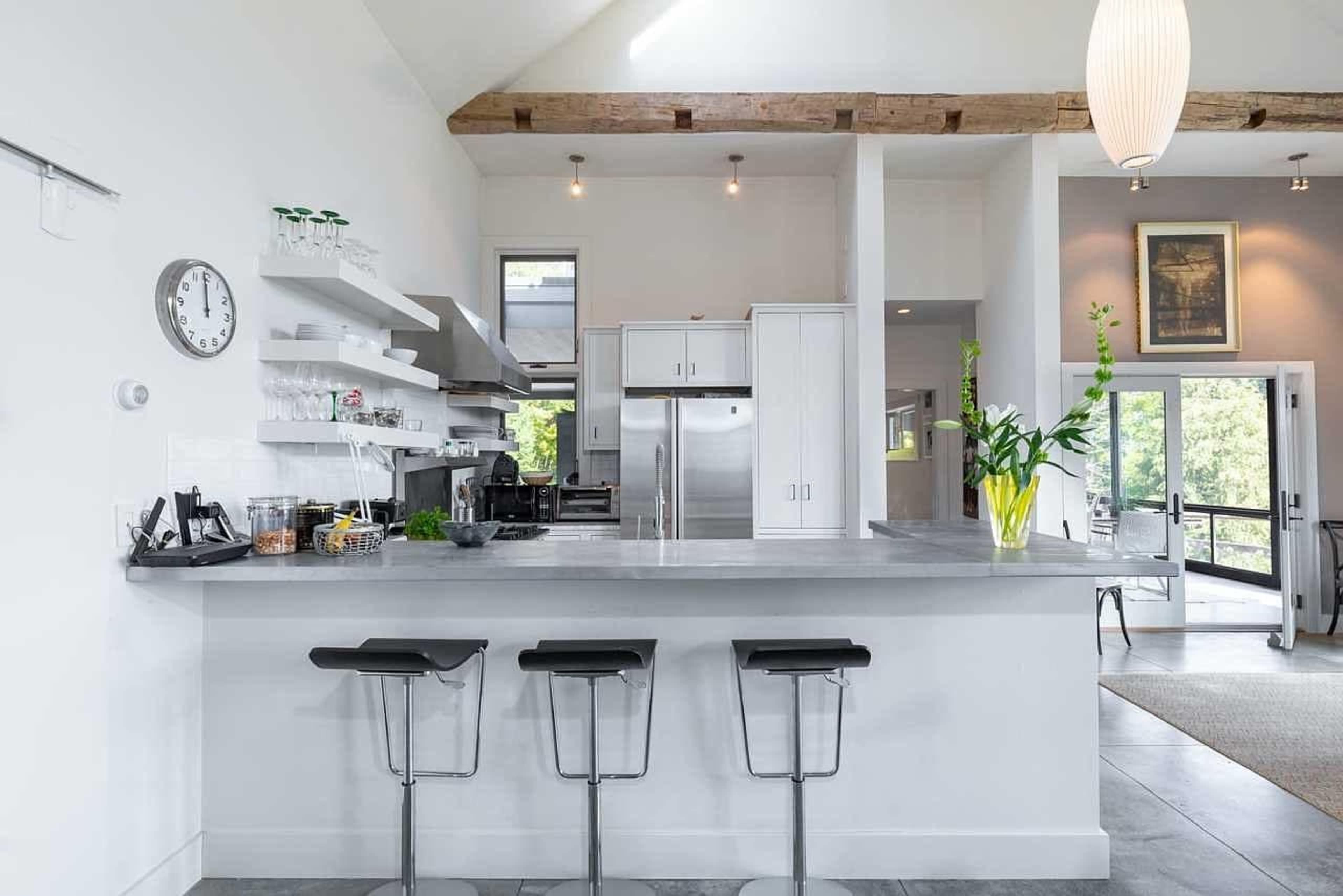 A modern kitchen features a long countertop with three bar stools, open shelving, and stainless steel appliances, all under a high ceiling with wooden beams.