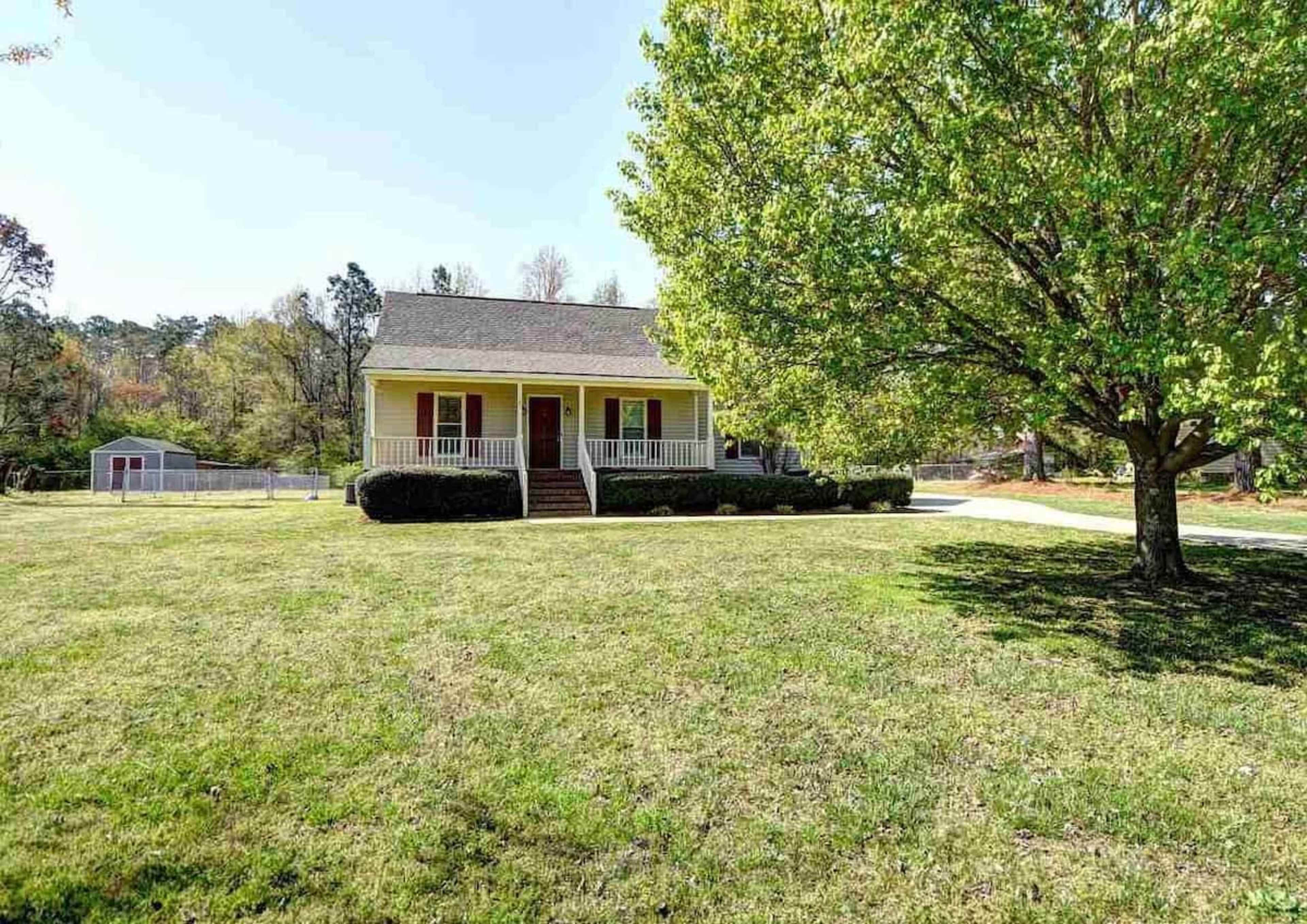A single-story yellow house with a front porch is situated on a grassy lawn, accompanied by a large tree nearby.