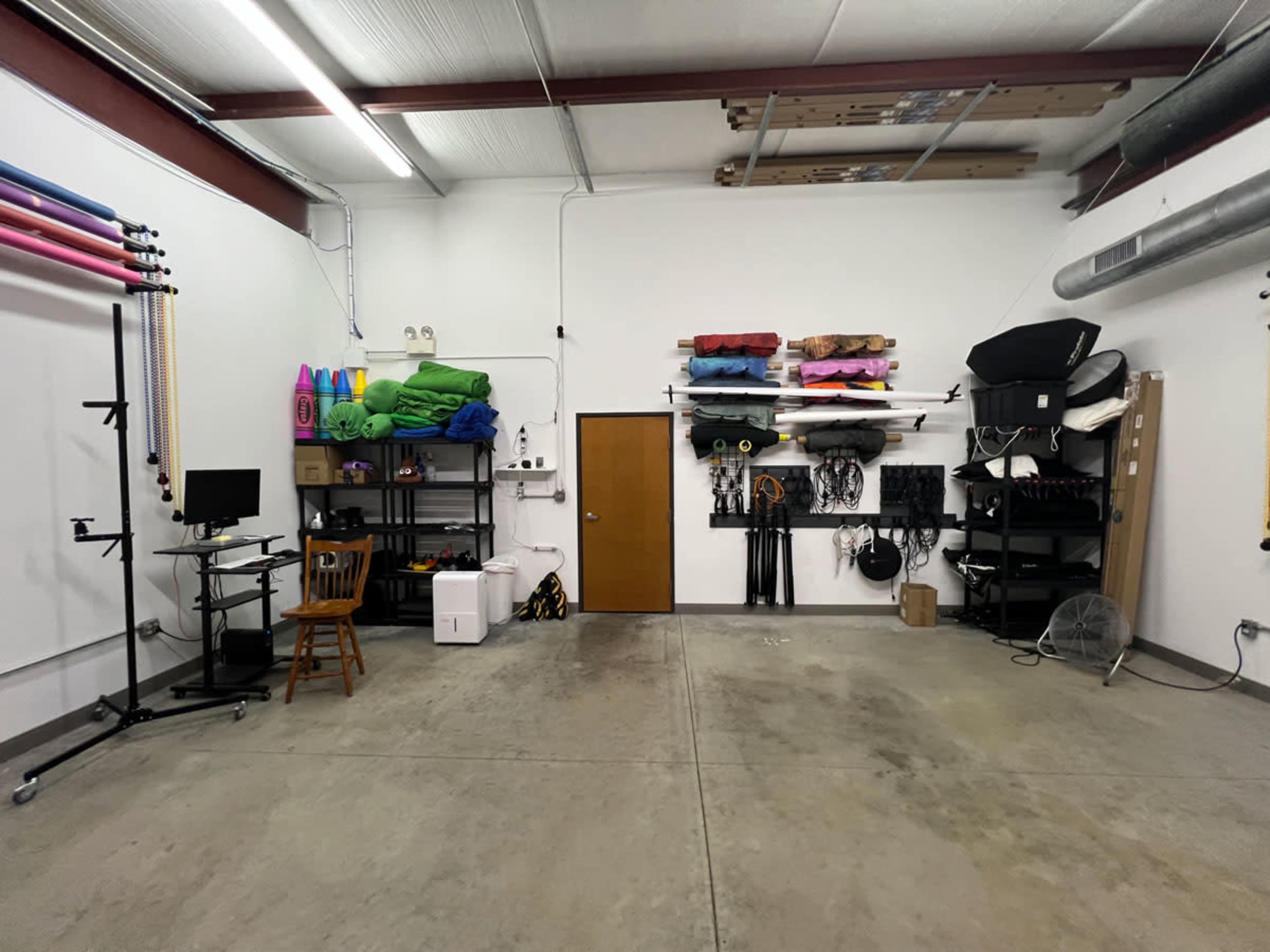 A spacious studio with a concrete floor, featuring shelves stocked with colorful equipment and props, a chair, and a door on one wall.