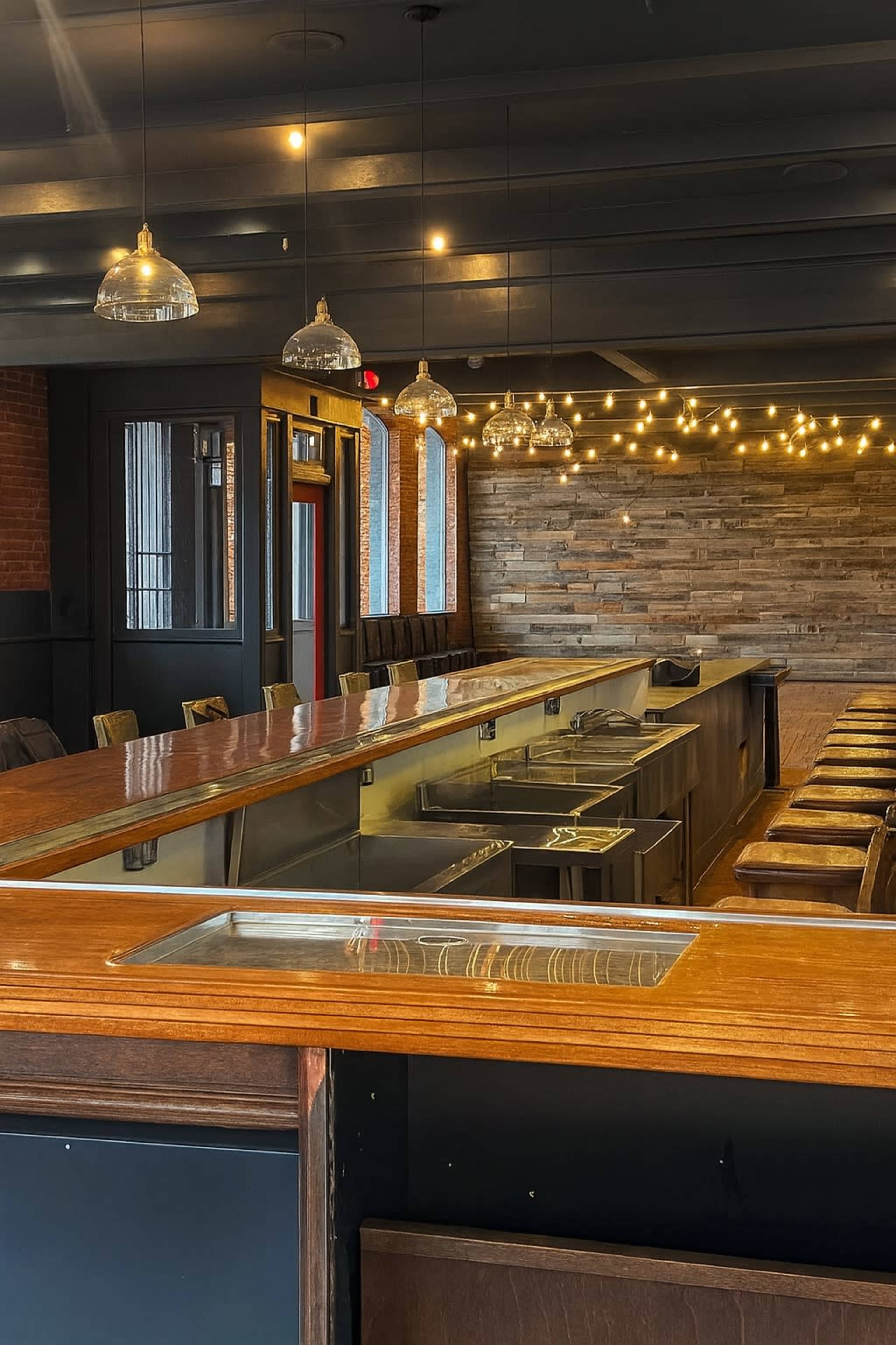 The image shows an empty restaurant bar with a wooden counter, metal fixtures, and pendant lighting, set against a backdrop of exposed brick and wooden wall paneling.