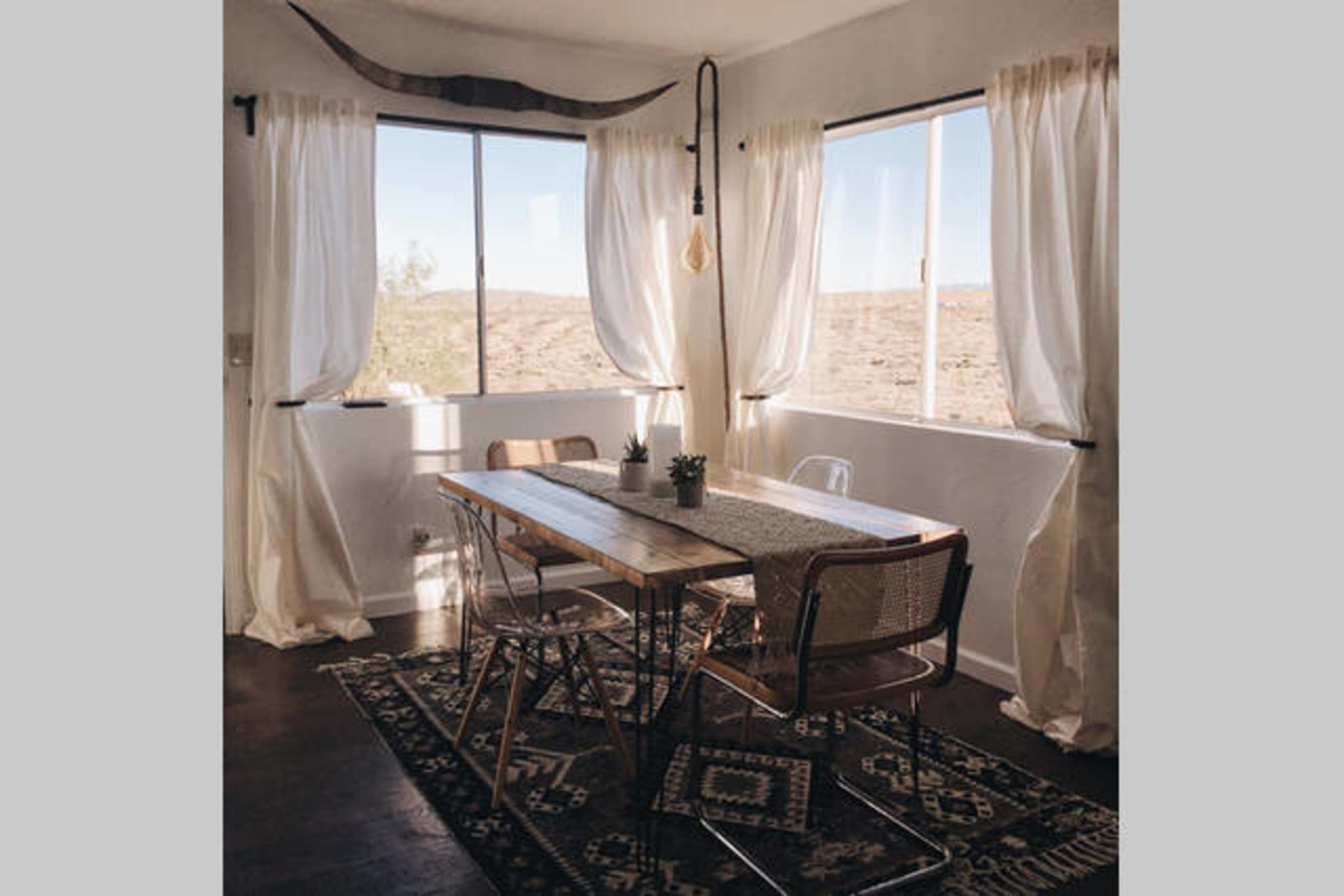 A kitchen dining area features a wooden table surrounded by metal and wicker chairs, with large windows and white drapes overlooking a desert landscape.