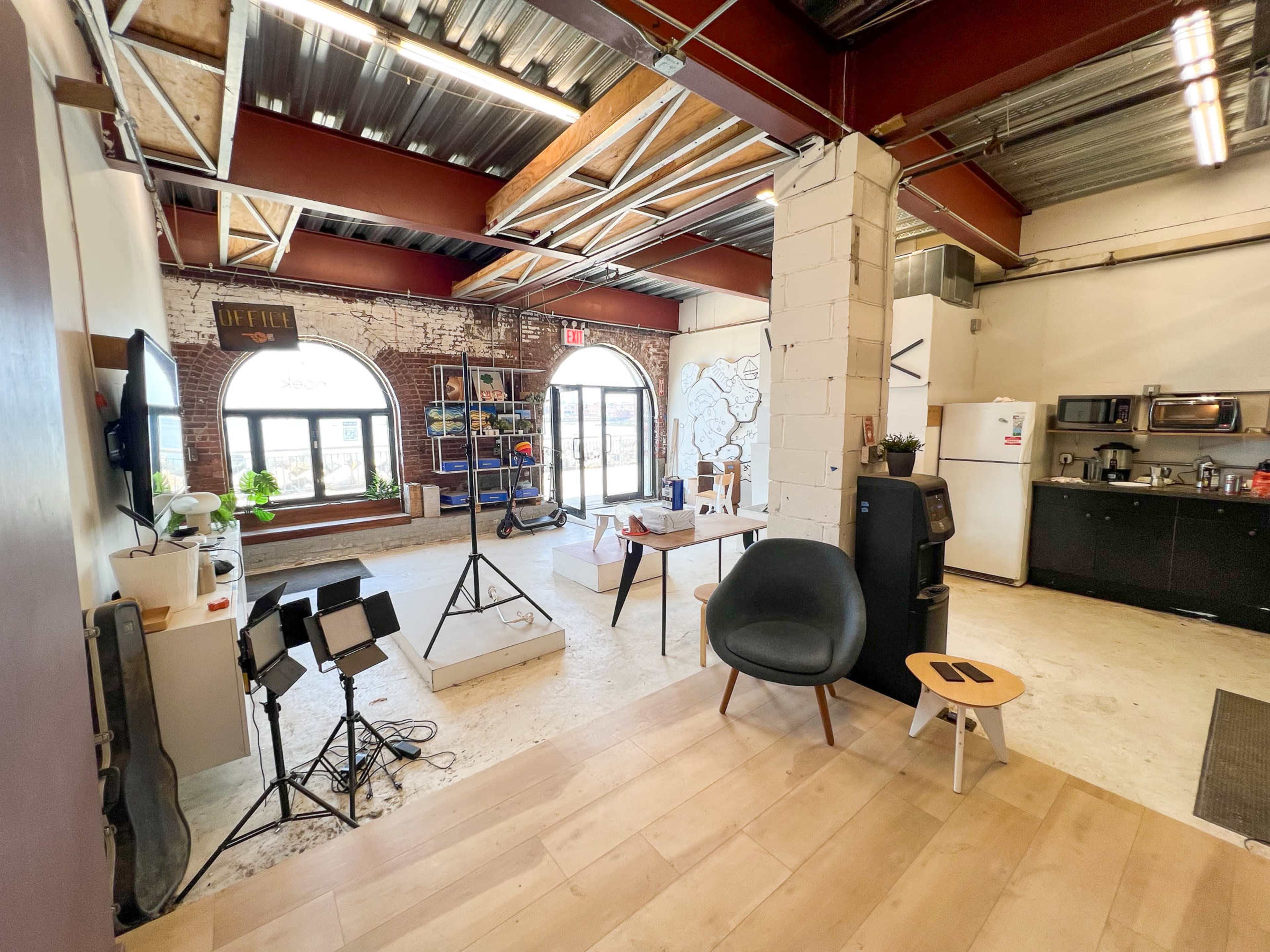 The image shows a modern office space with exposed brick walls, large windows, a wooden floor, and various furniture items including a chair, a table, and a small kitchen area.