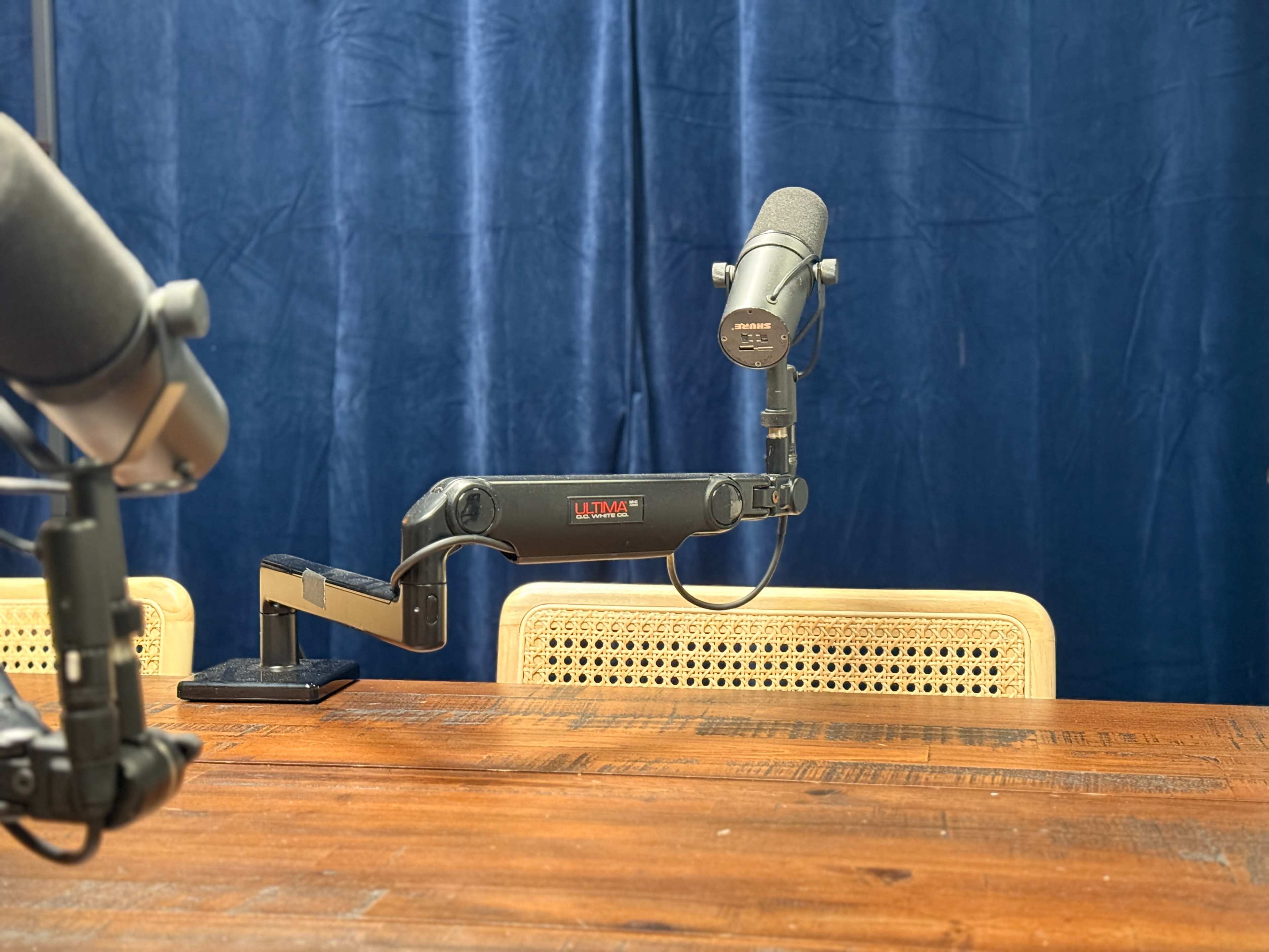 The image shows a wooden table with two microphone stands attached to an arm, positioned in front of a blue curtain.