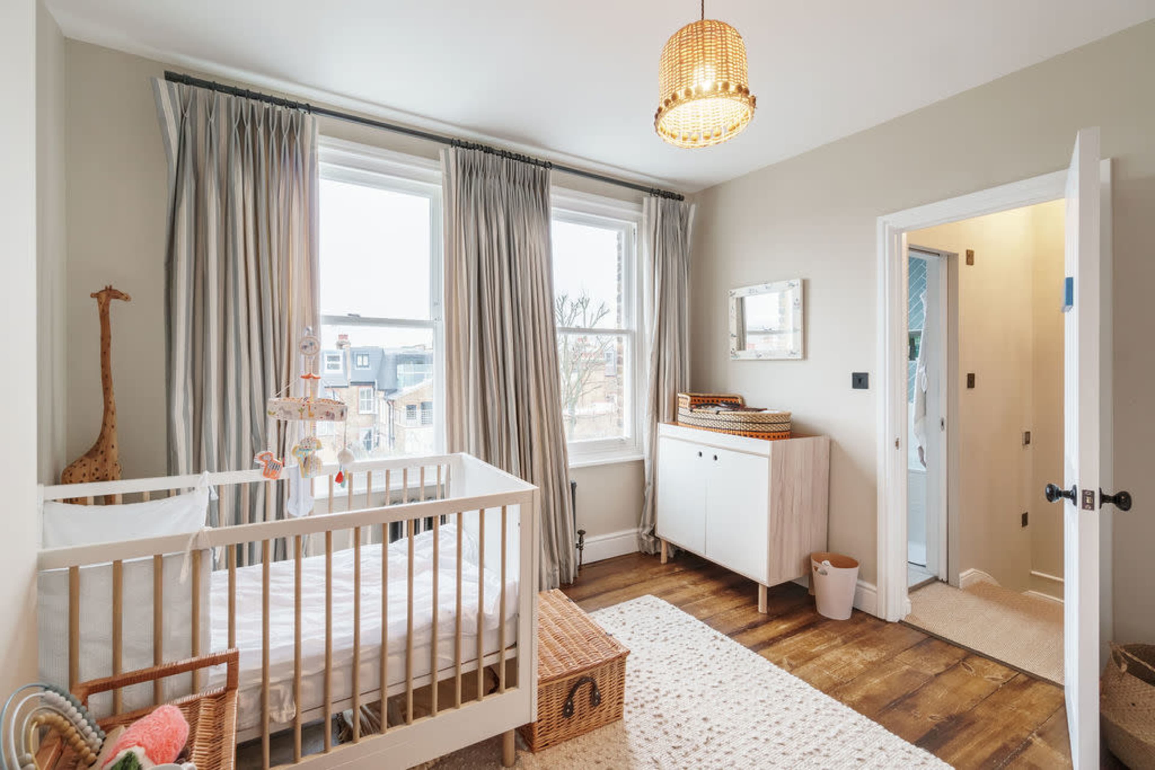 A softly lit nursery featuring a crib next to large windows with curtains, a dresser, and a door leading to another room.