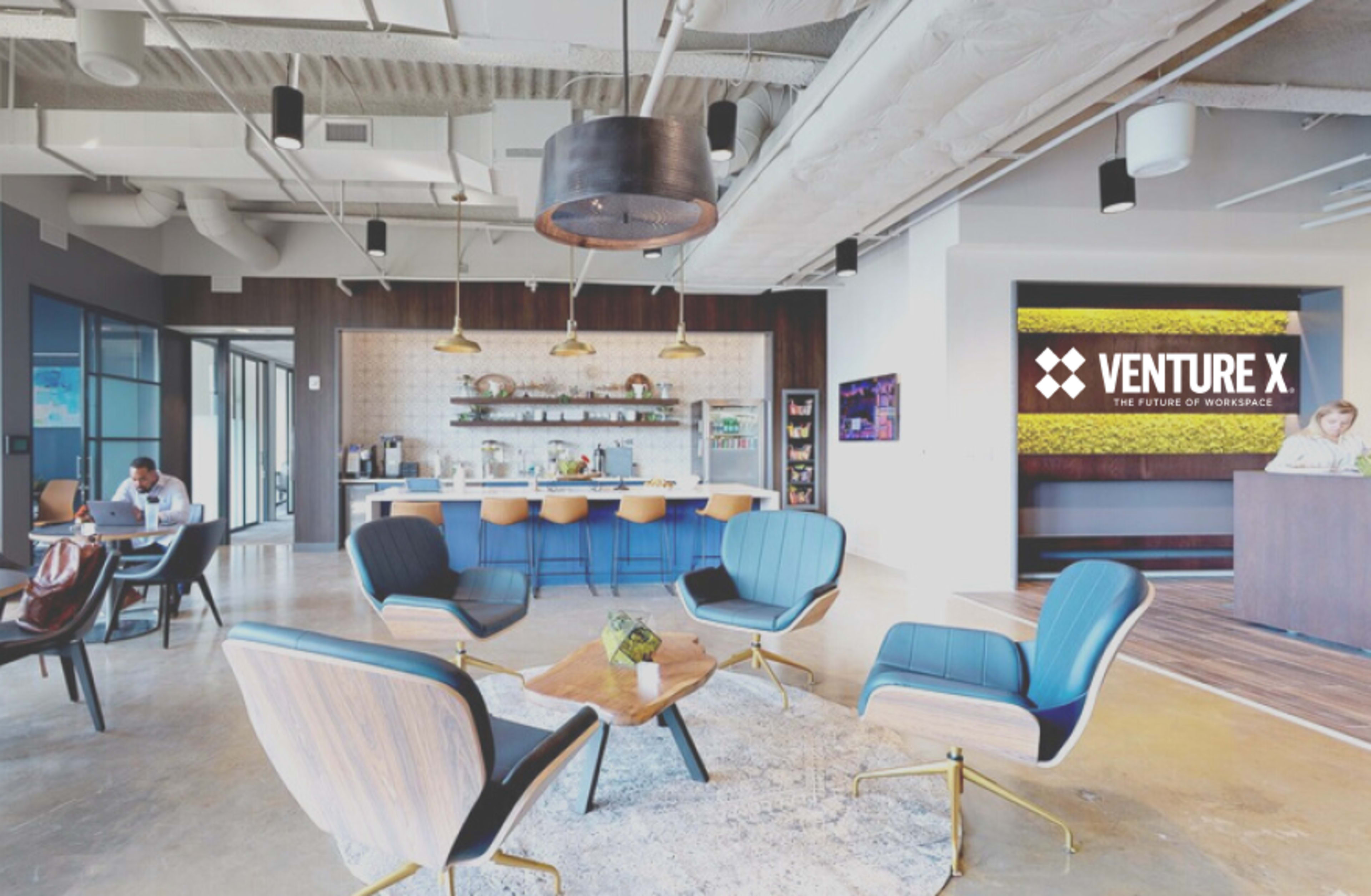 A modern office space features a lounge area with blue chairs, a central table, and a kitchenette in the background, along with a reception desk displaying a logo for Venture X.