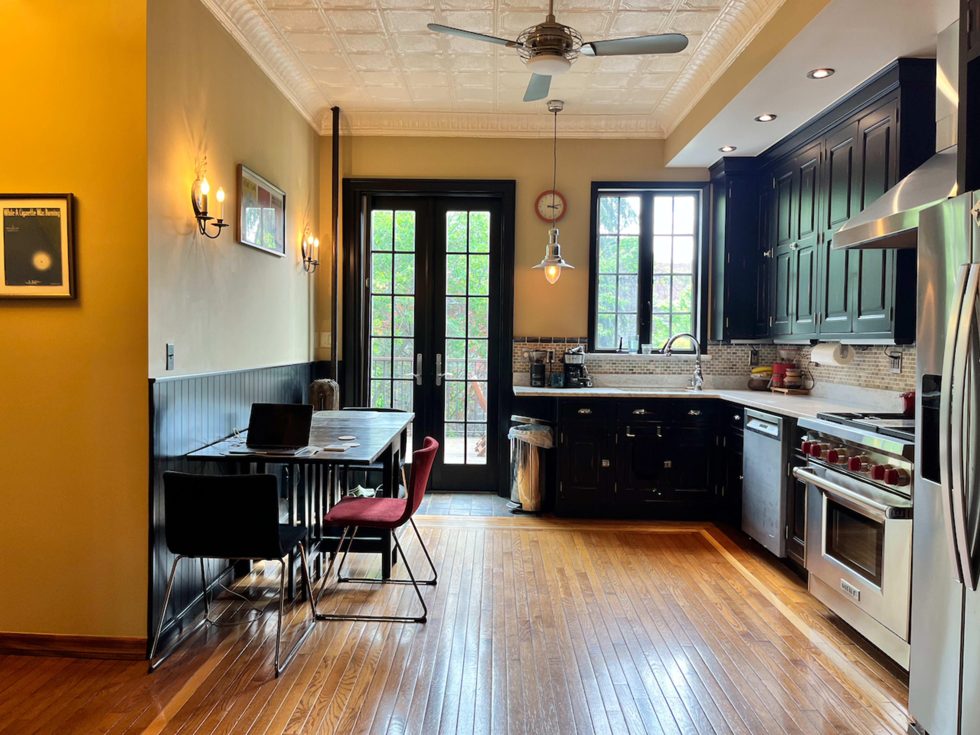 A kitchen with dark cabinets, stainless steel appliances, a dining table with black and red chairs, and large windows revealing a view of greenery outside.