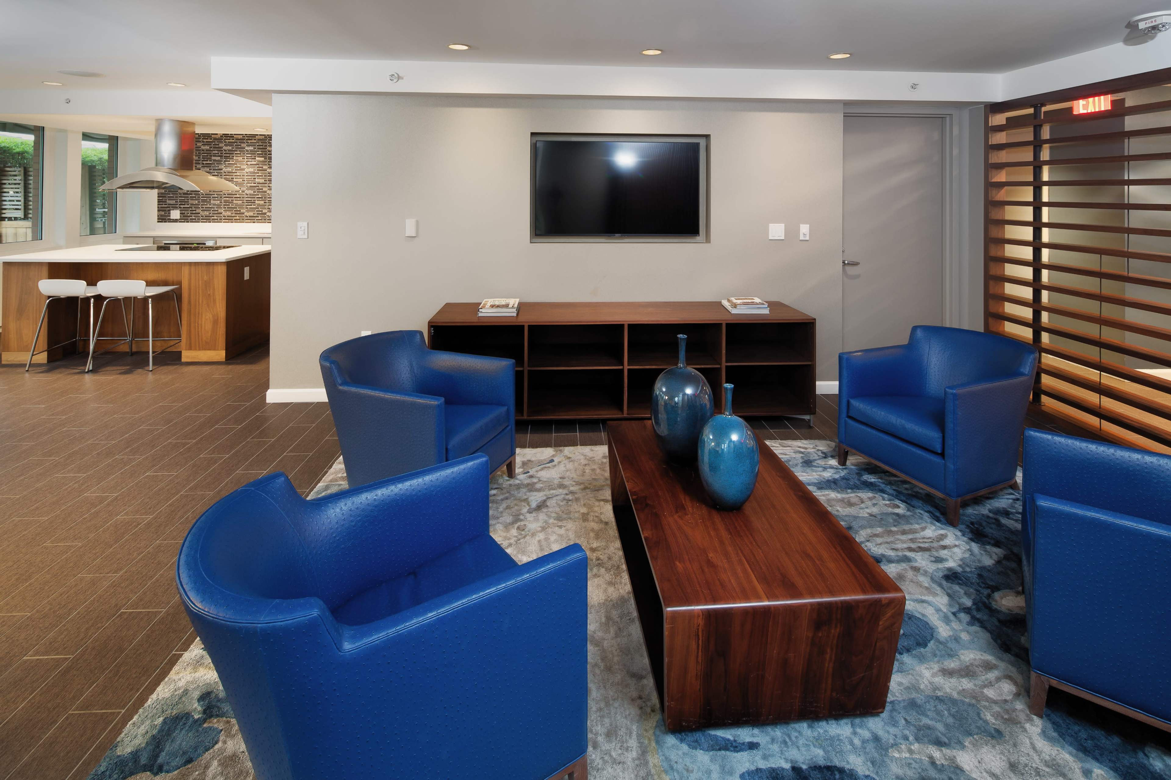 A modern lounge area features four blue chairs surrounding a wooden coffee table, with a flat-screen TV mounted on the wall and an open kitchen visible in the background.