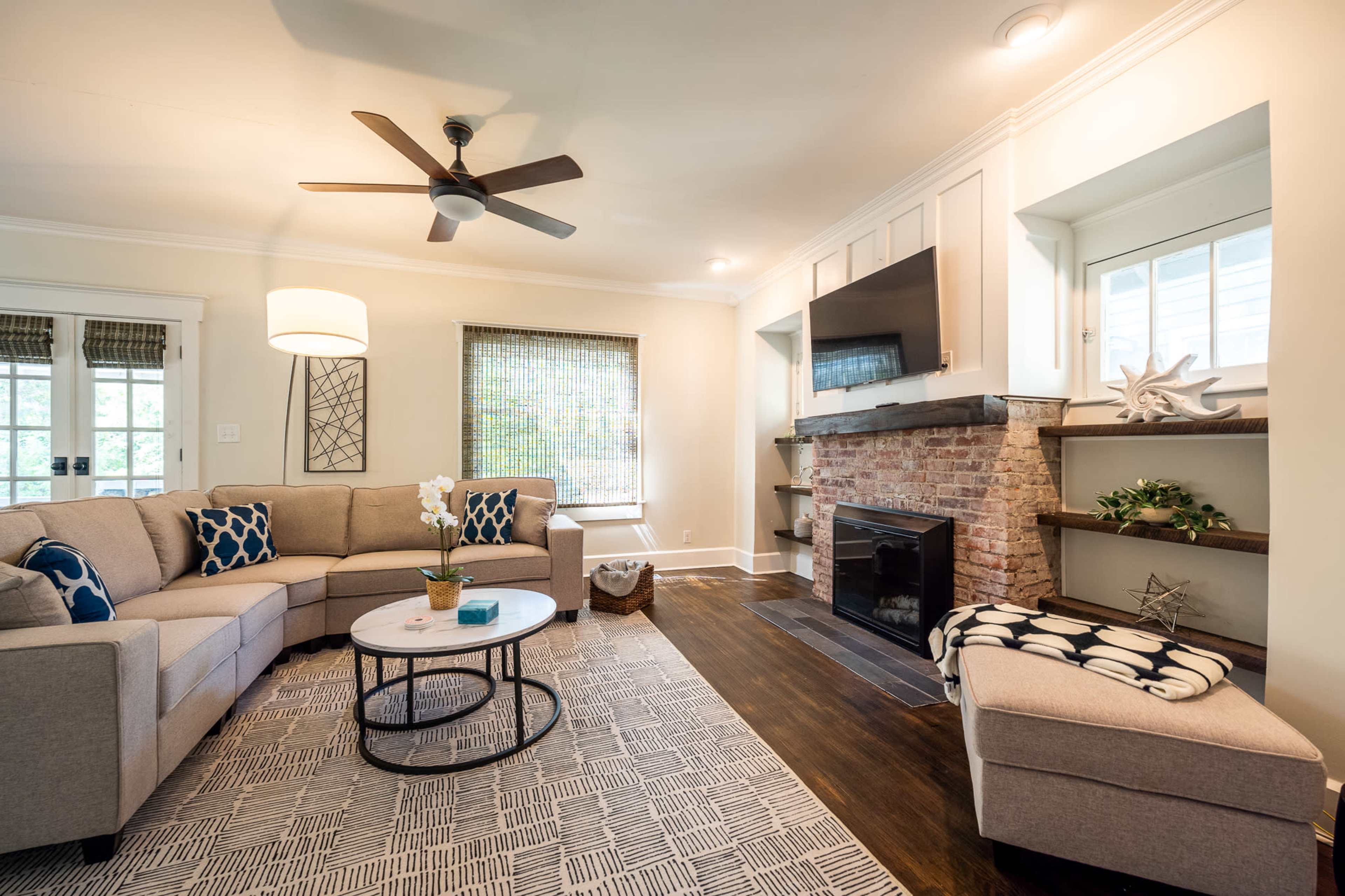 A modern living room features a sectional sofa, a round coffee table, a flat-screen TV mounted above a brick fireplace, and a light fixture, all complemented by a patterned area rug and decorative shelves.