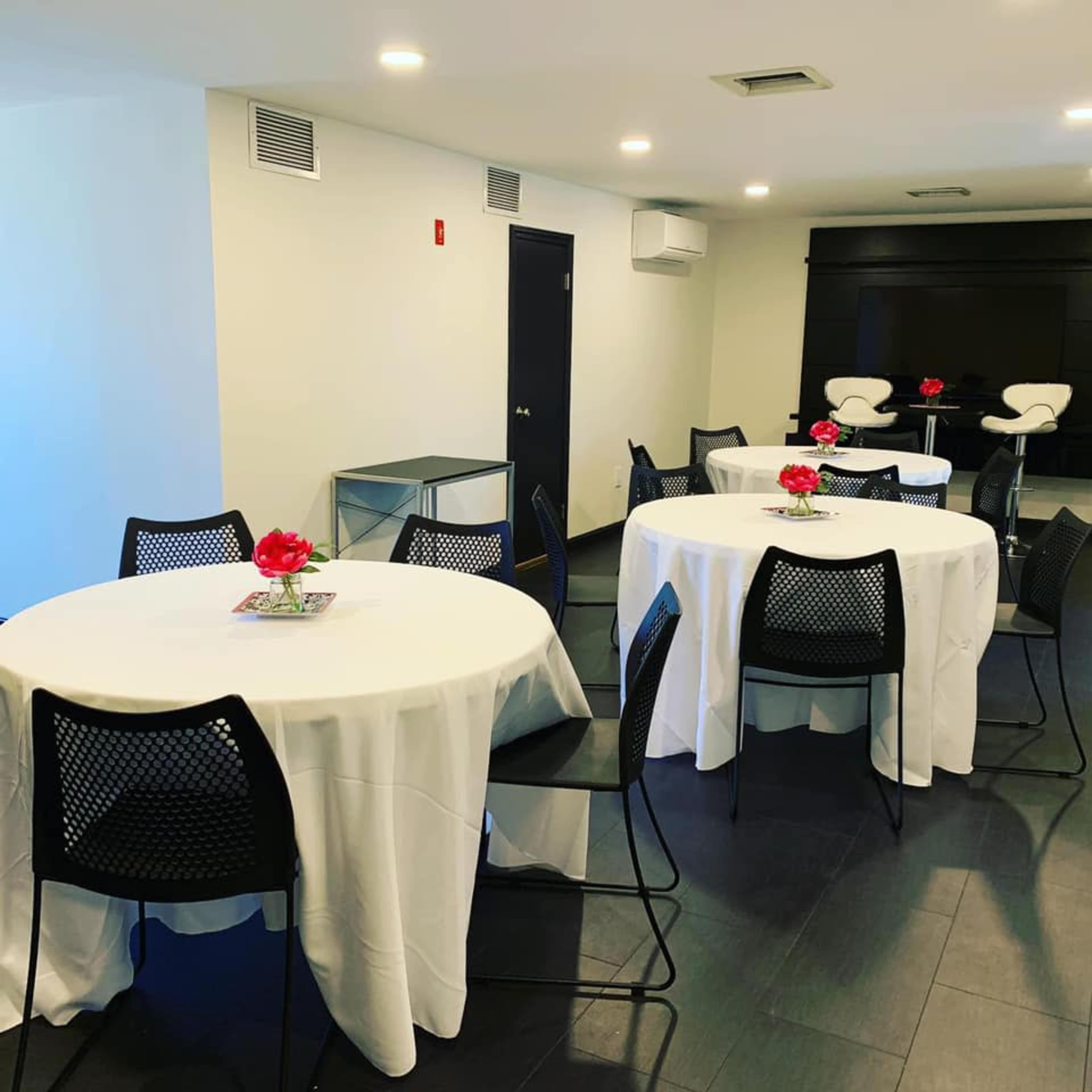 The image shows a modern meeting room with round tables covered in white tablecloths, each adorned with a small flower arrangement.