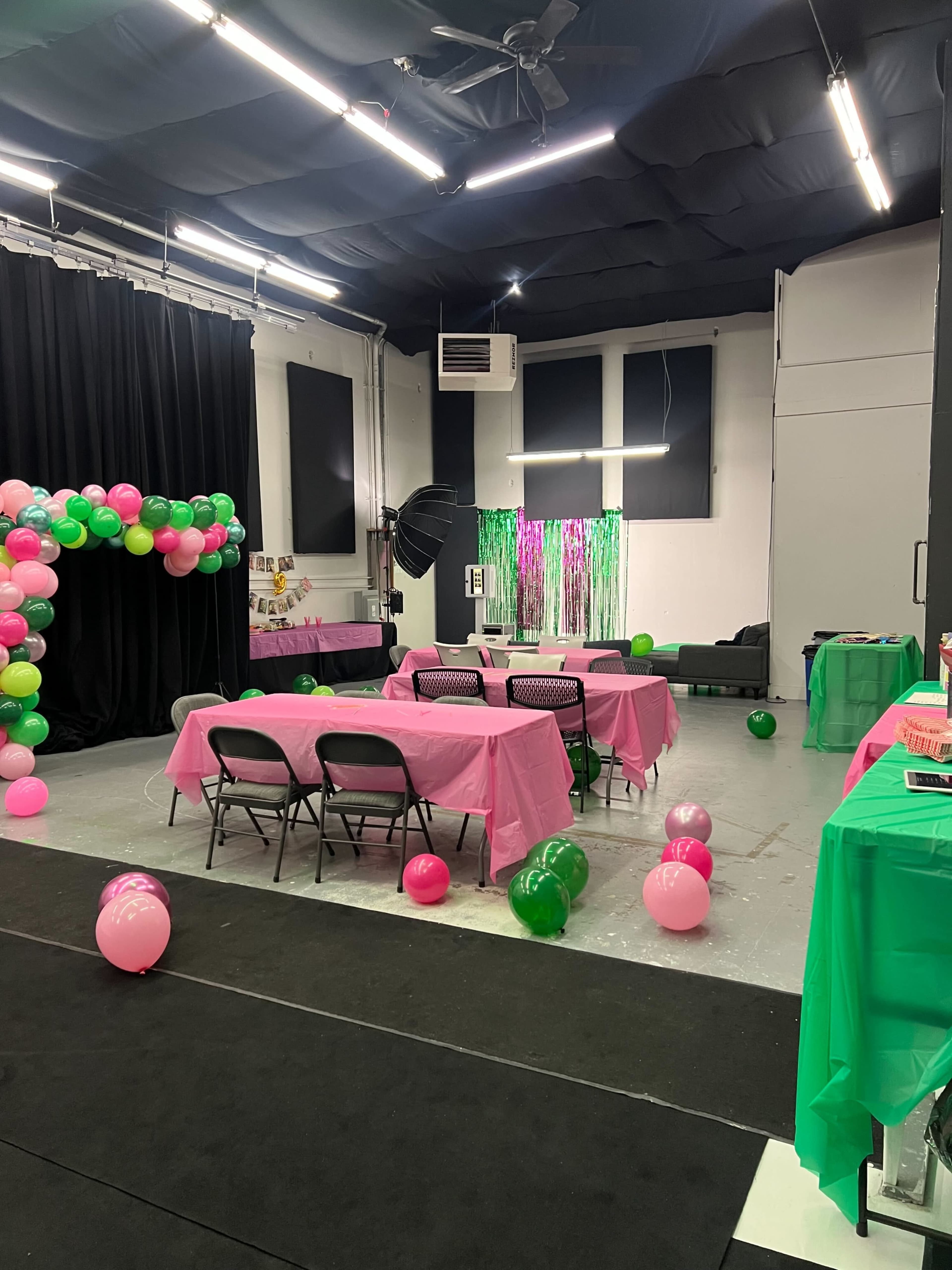 The image shows a party setup in a studio space with pink and green decorations, tables covered in pink tablecloths, and colorful balloons scattered around.