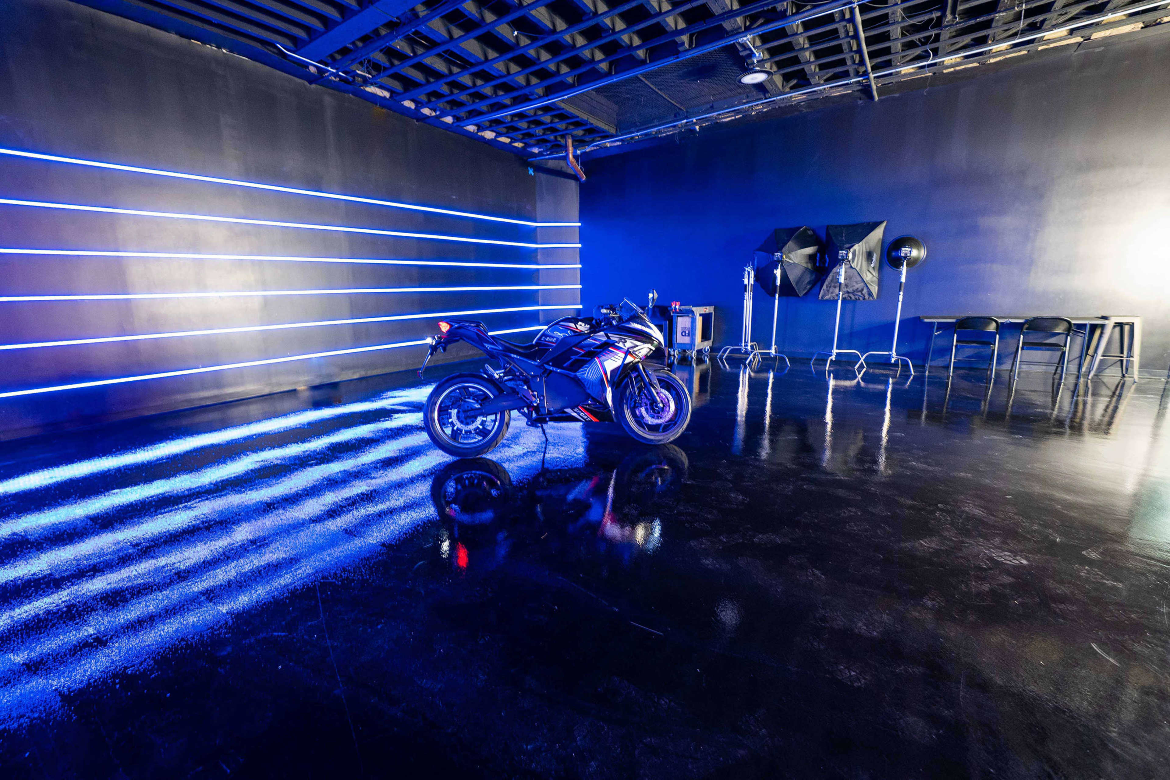 A motorcycle is positioned in a sleek, modern studio space illuminated by blue neon lights and featuring a reflective floor.