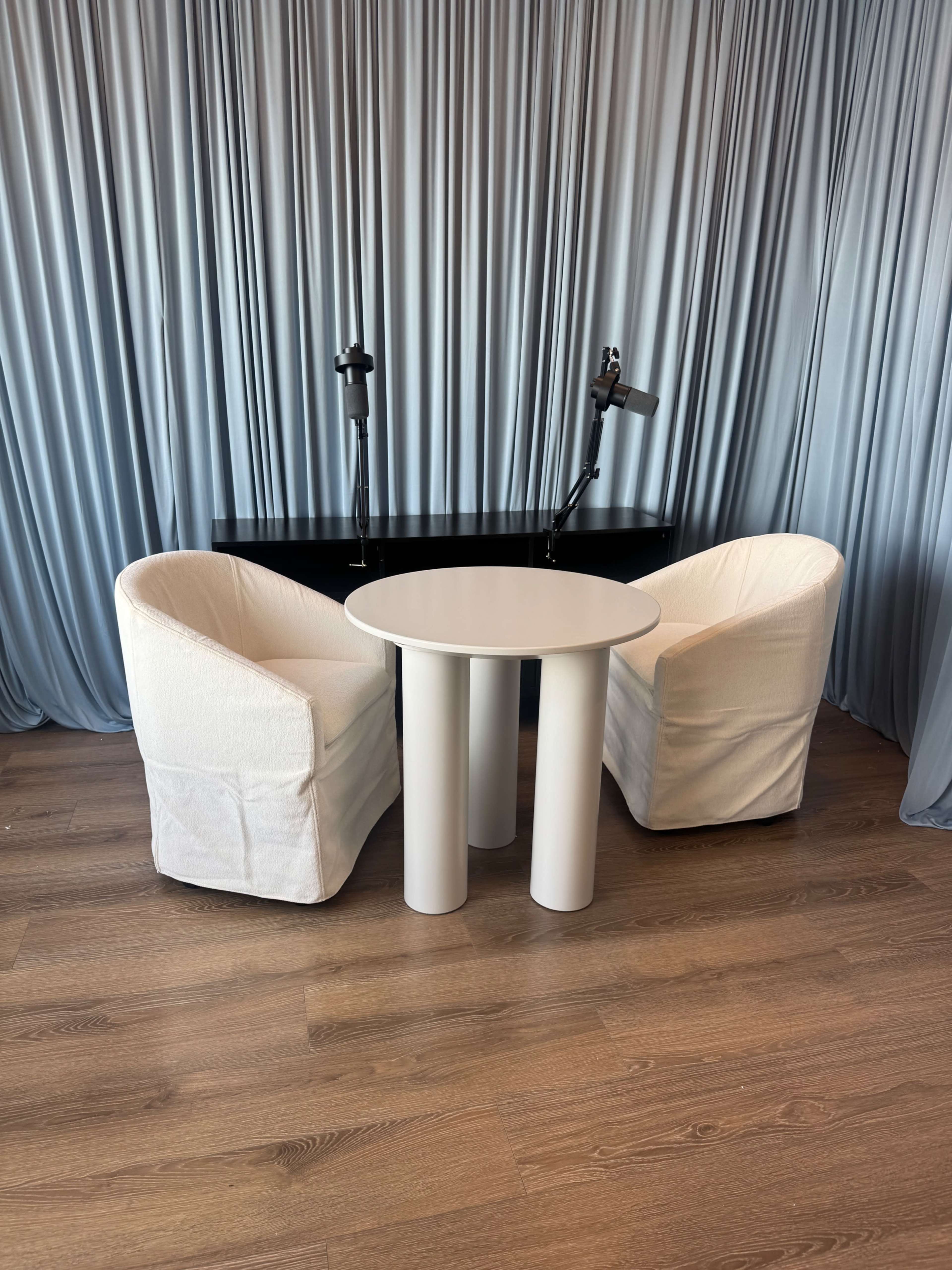 The image shows two white upholstered chairs positioned around a round white table in front of a backdrop of draped fabric.