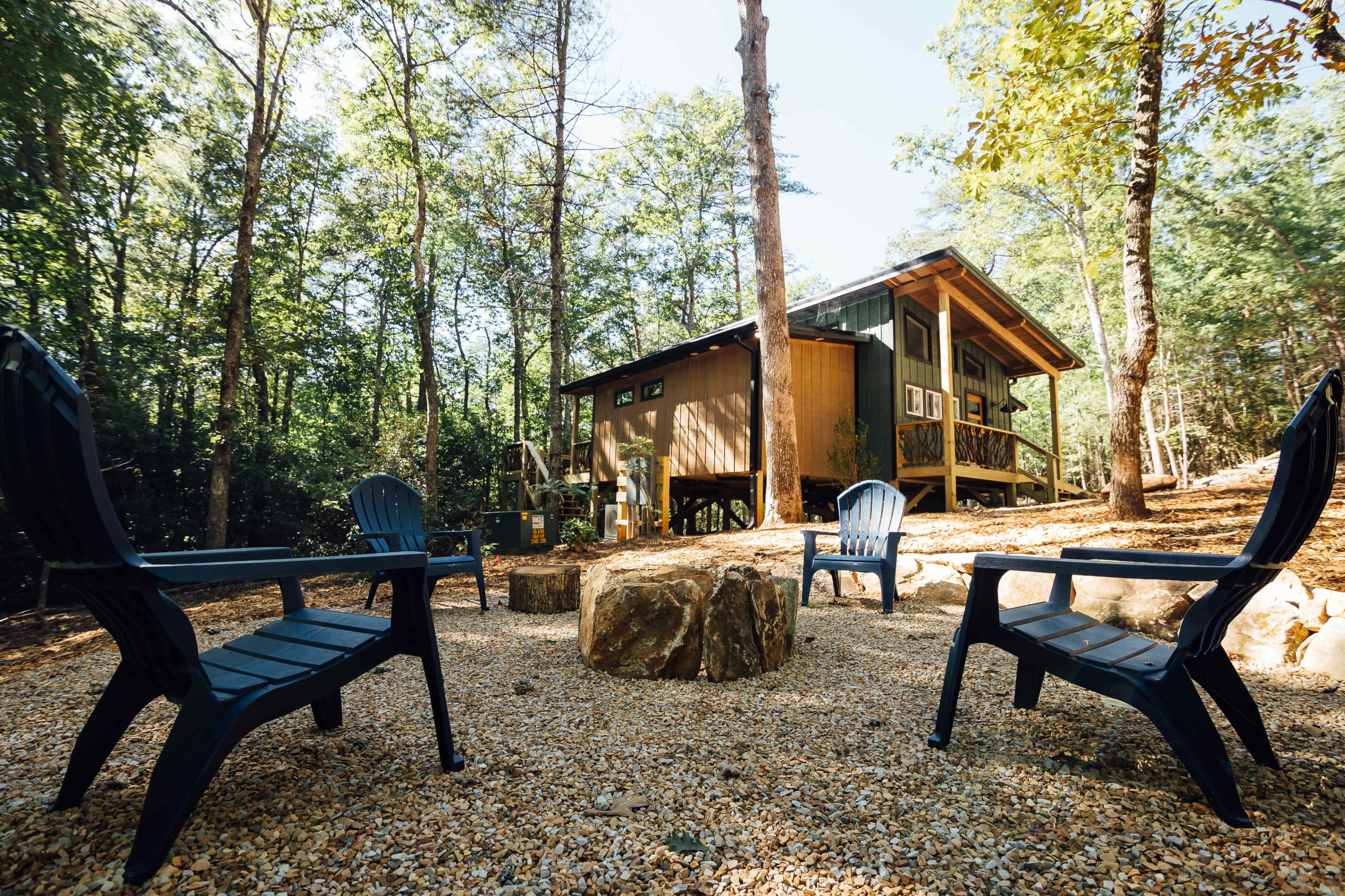 A modern cabin set among trees with four outdoor chairs arranged around a large rock in a gravel area.