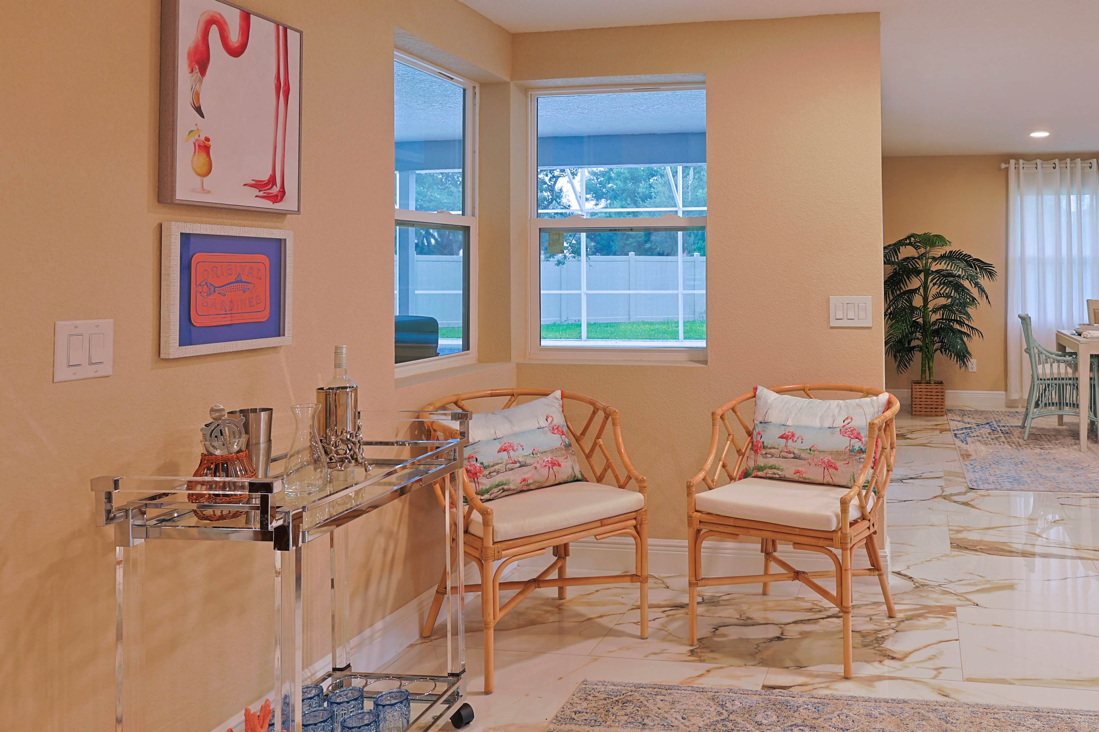 The image shows a cozy sitting area with two rattan chairs, a side table, and a bar cart against a wall adorned with artwork and windows letting in natural light.