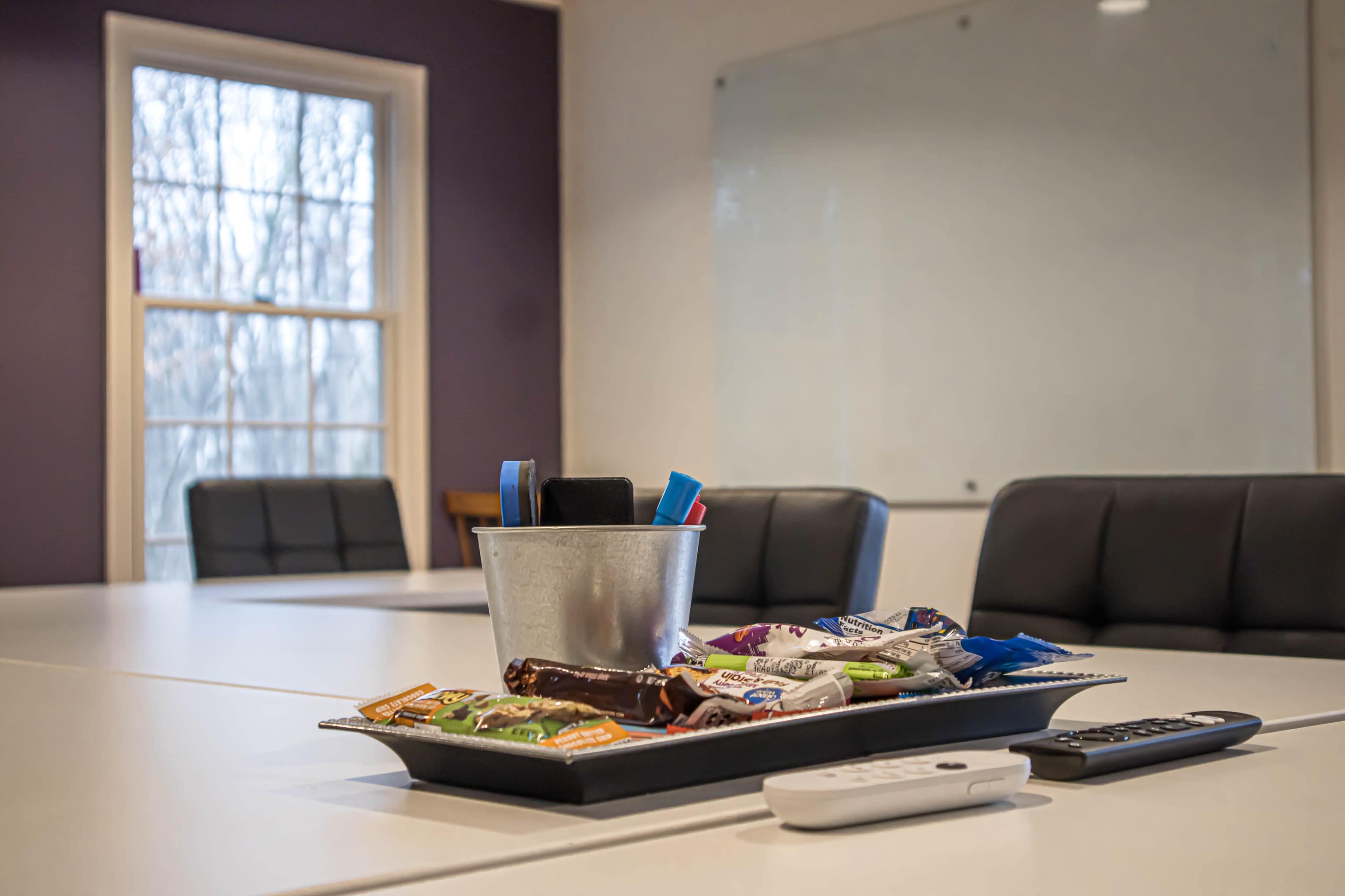 A conference room table is set with a tray of snacks and writing utensils, with chairs and a whiteboard visible in the background.