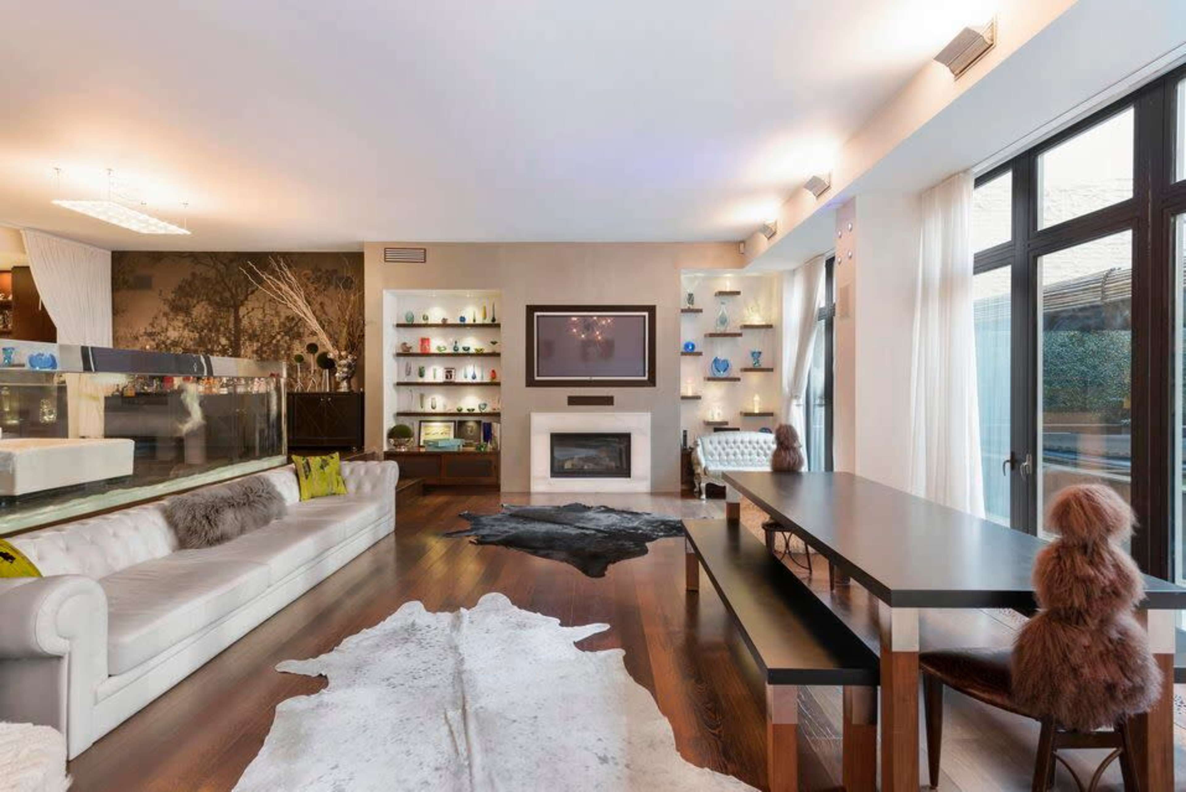 A spacious modern living room featuring a white leather sofa, a dark wooden dining table, and decorative shelves with various objects.
