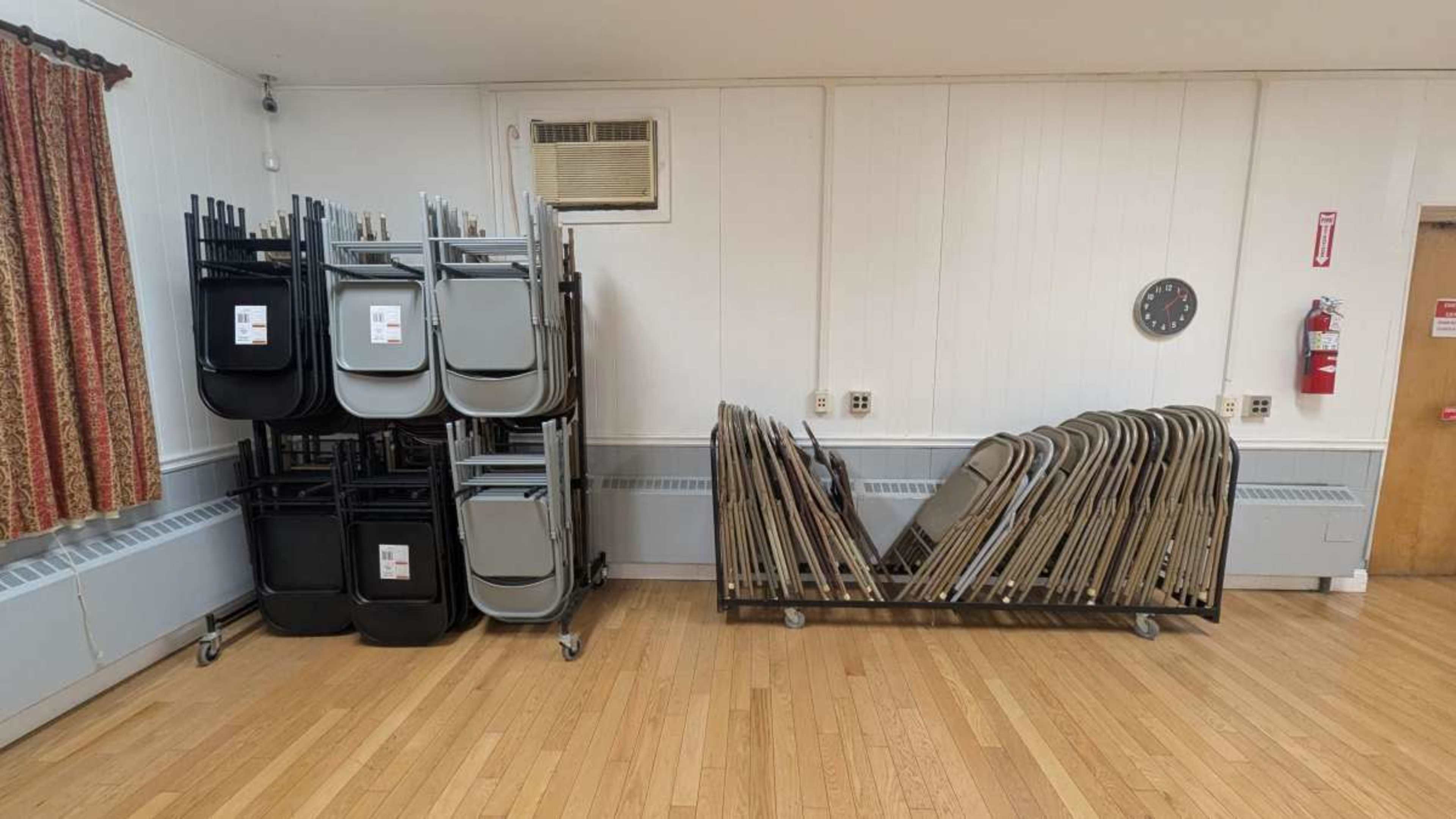 The image shows a room with a stack of folding chairs on the left and a rolled rack of folding tables on the right, with a clock and a fire extinguisher on the wall.