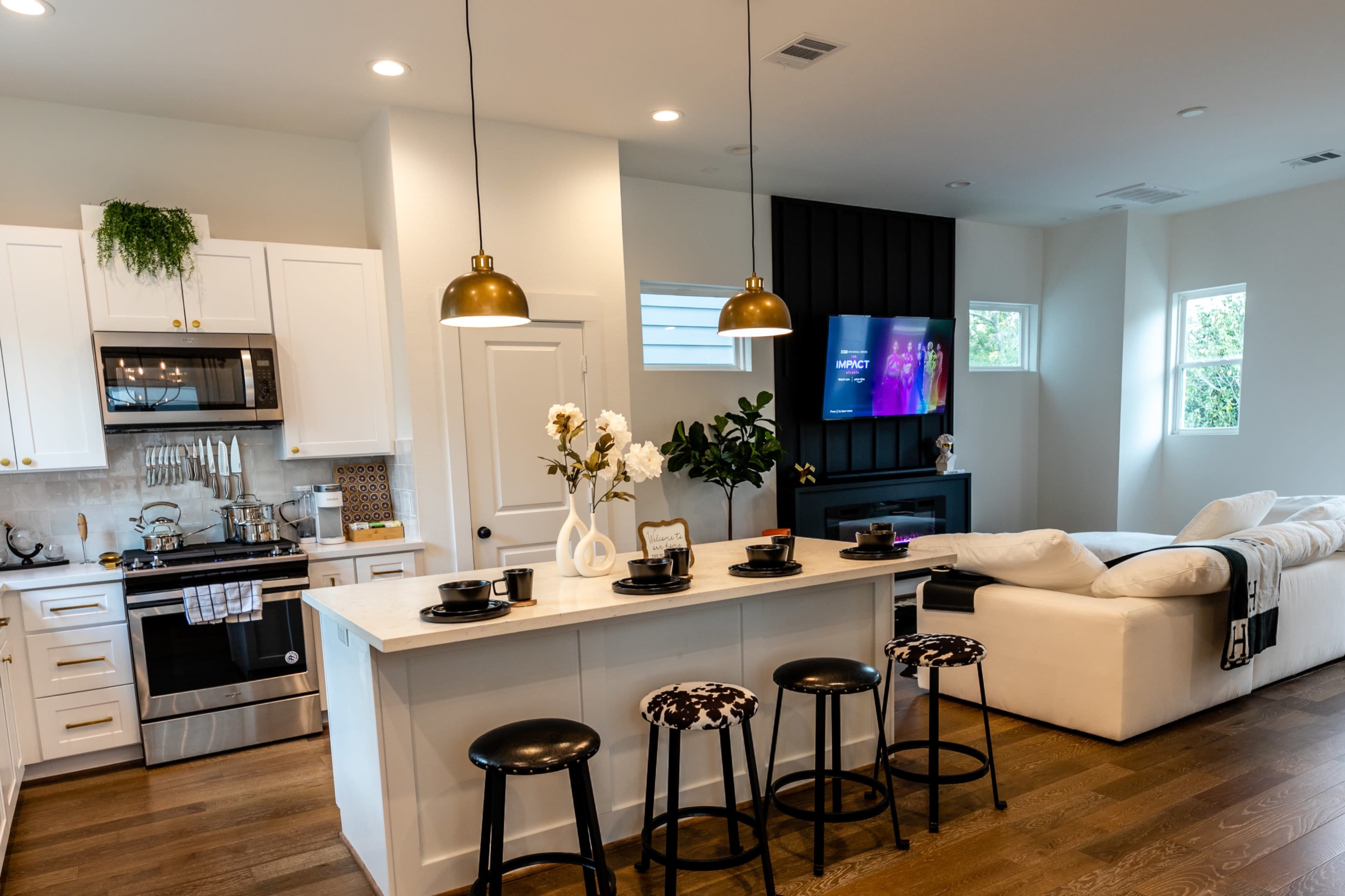 A modern kitchen and living area, featuring white cabinets, a bar with stools, and a couch in a light-filled space.