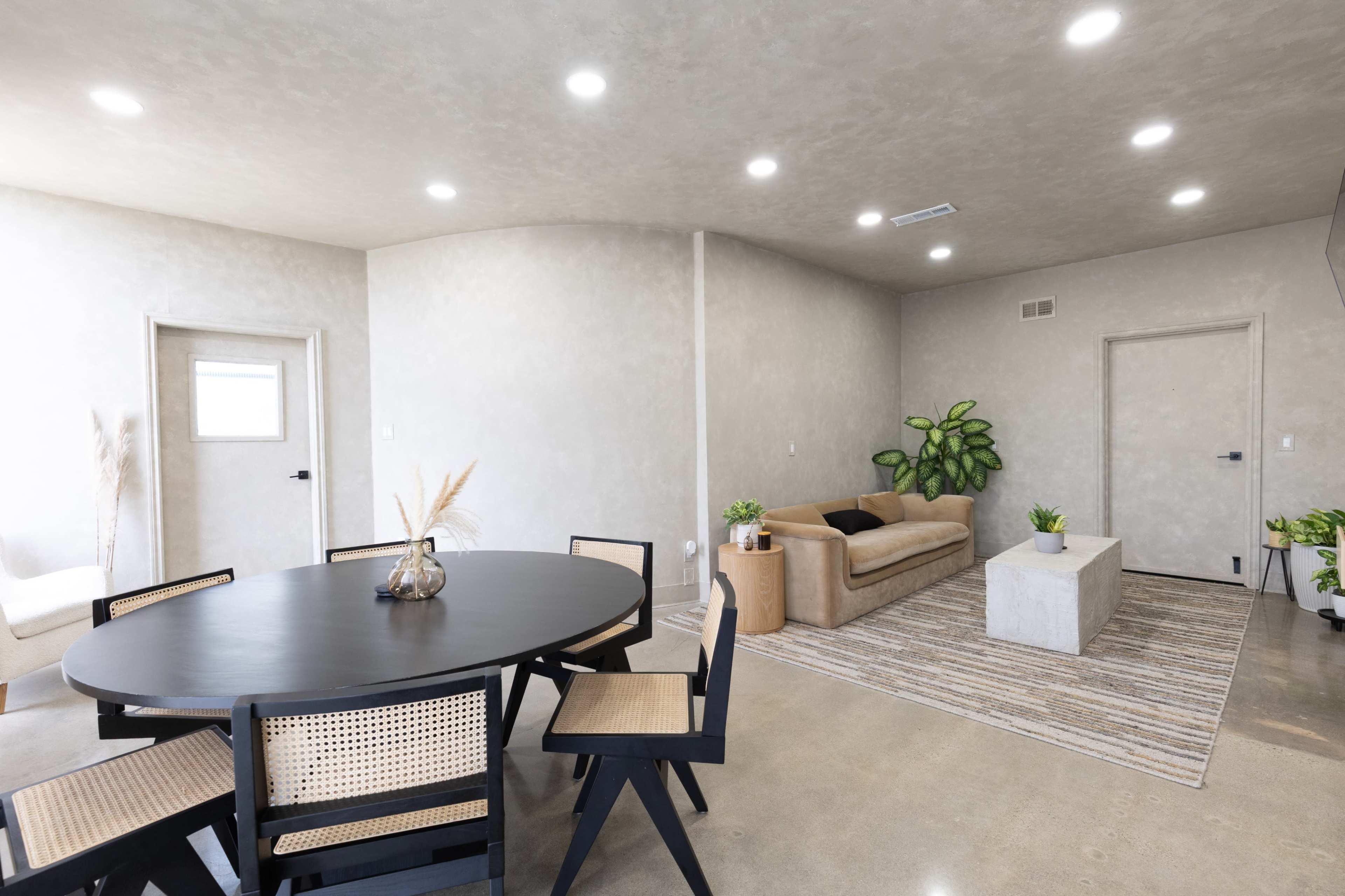 The image shows a modern living space with a round dining table and chairs, a beige sofa, a decorative coffee table, and various green plants.