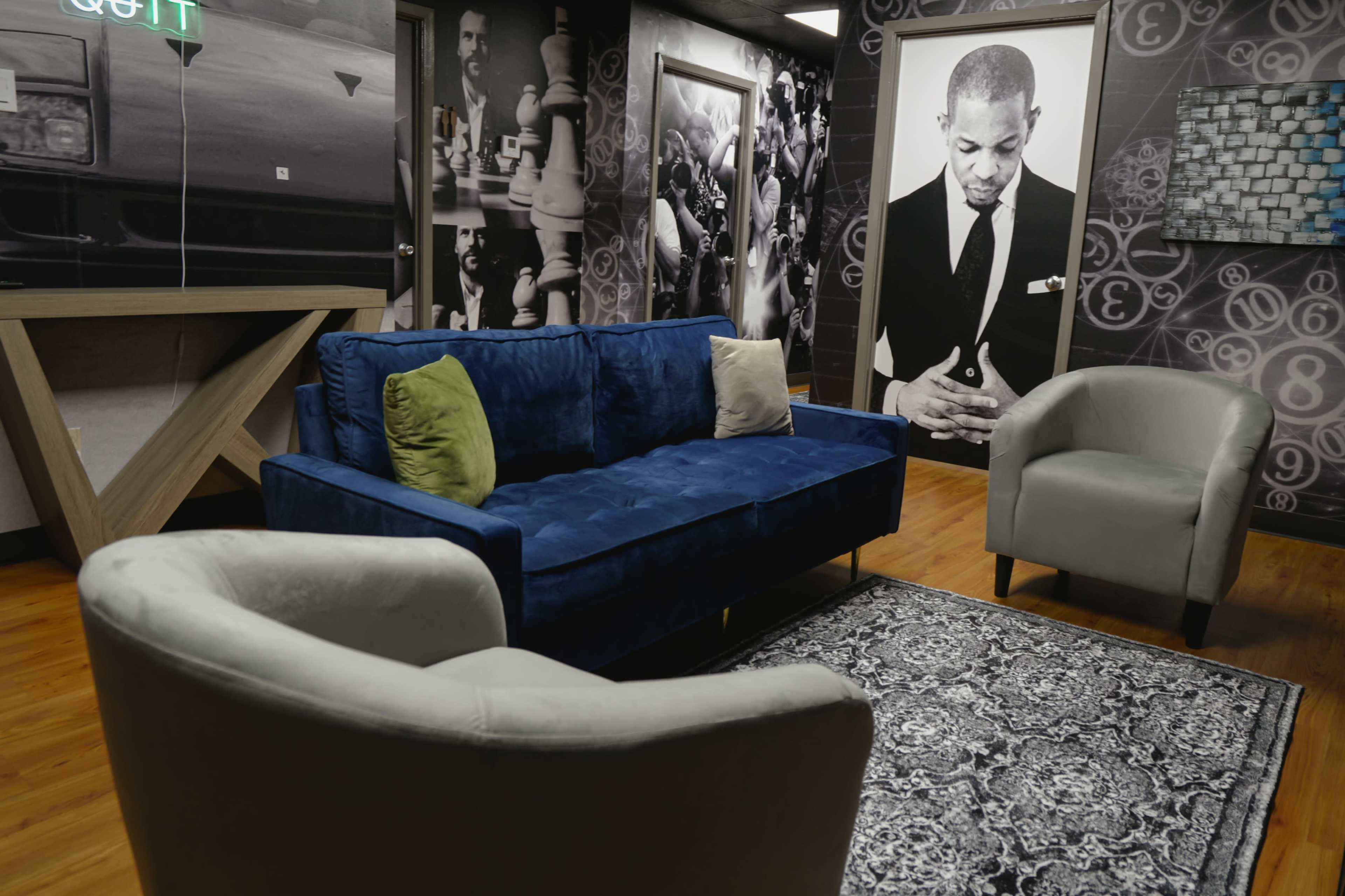The image features a modern lounge area with a blue couch, two light gray armchairs, and a decorative rug, set against walls adorned with black and white photographs.