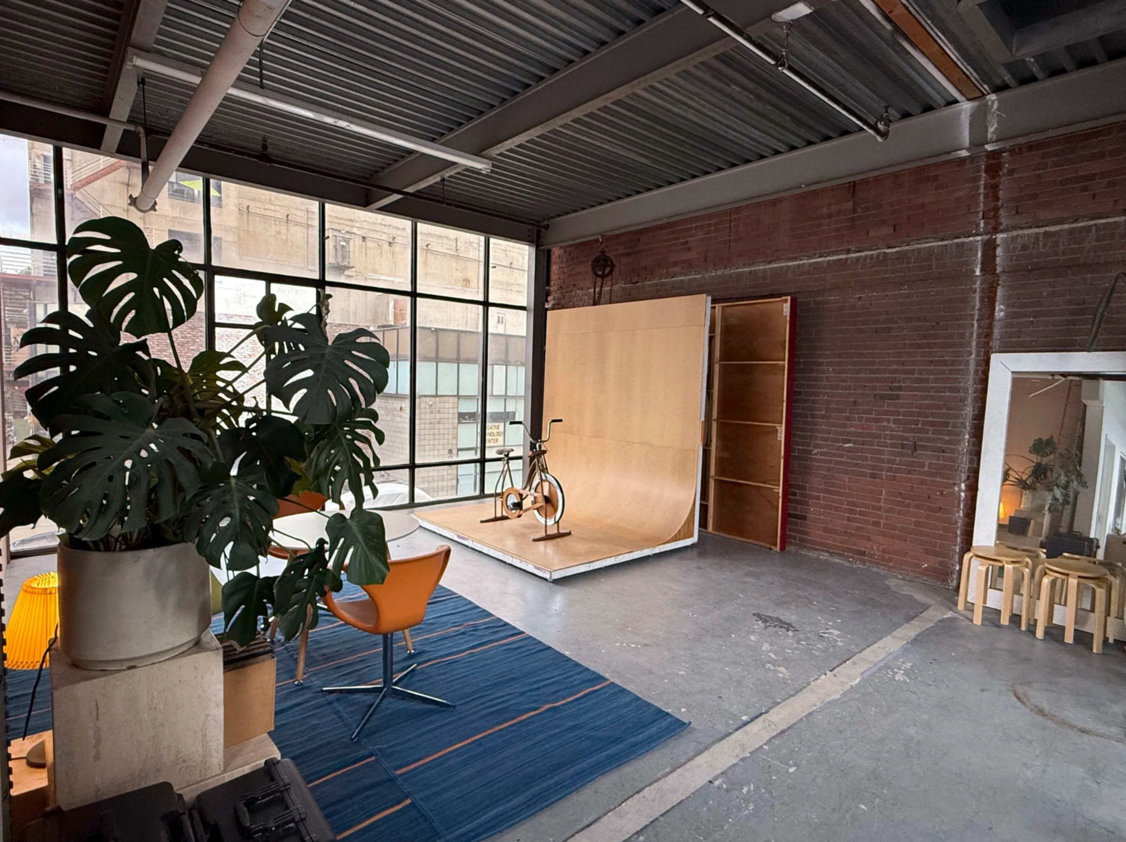 The image shows an industrial-style room with large windows, featuring a wooden ramp, a bicycle, a table with chairs, and a plant.