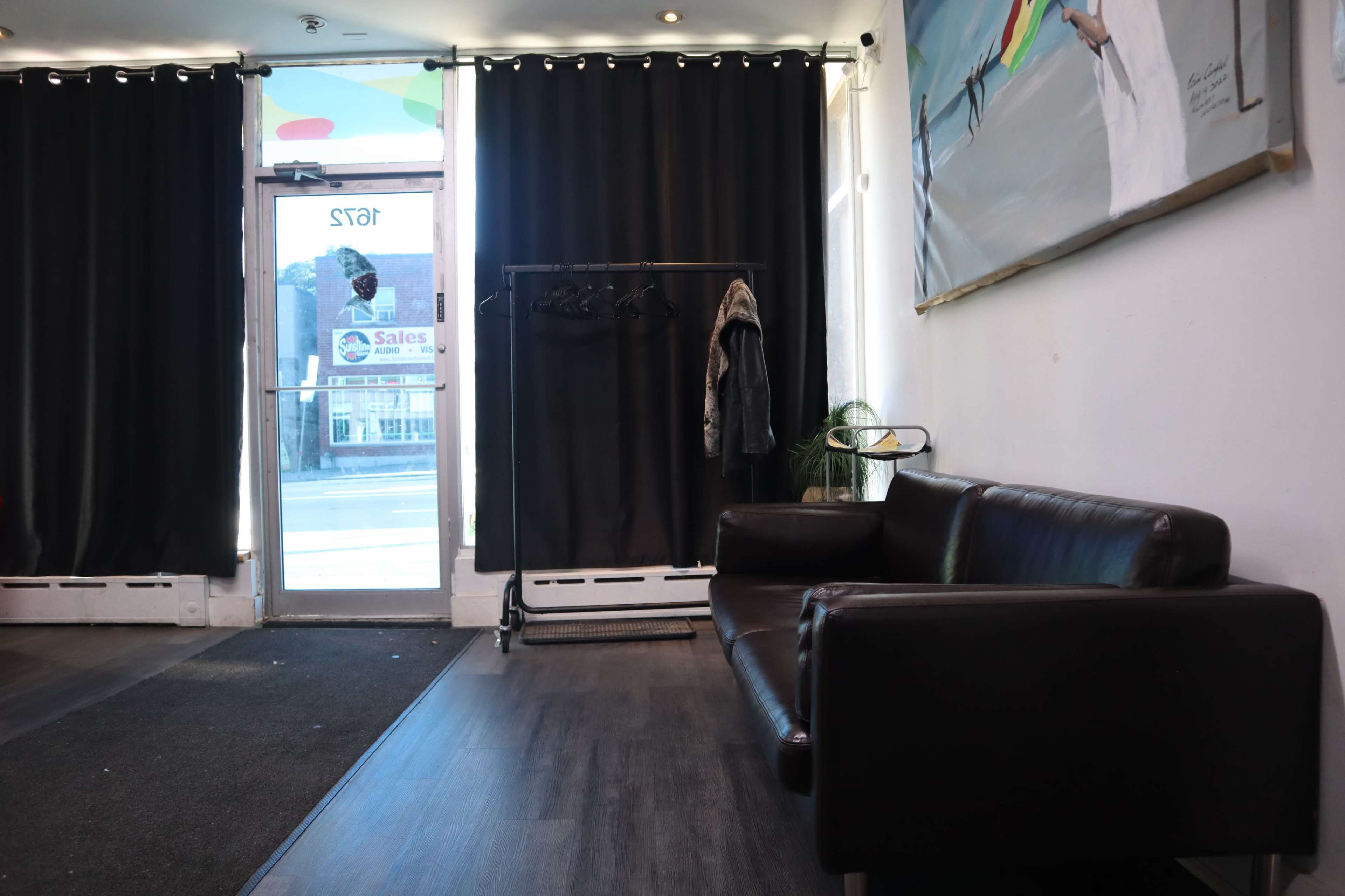 The image shows a waiting area with a dark brown couch, a clothing rack, and large black curtains beside a front glass door.