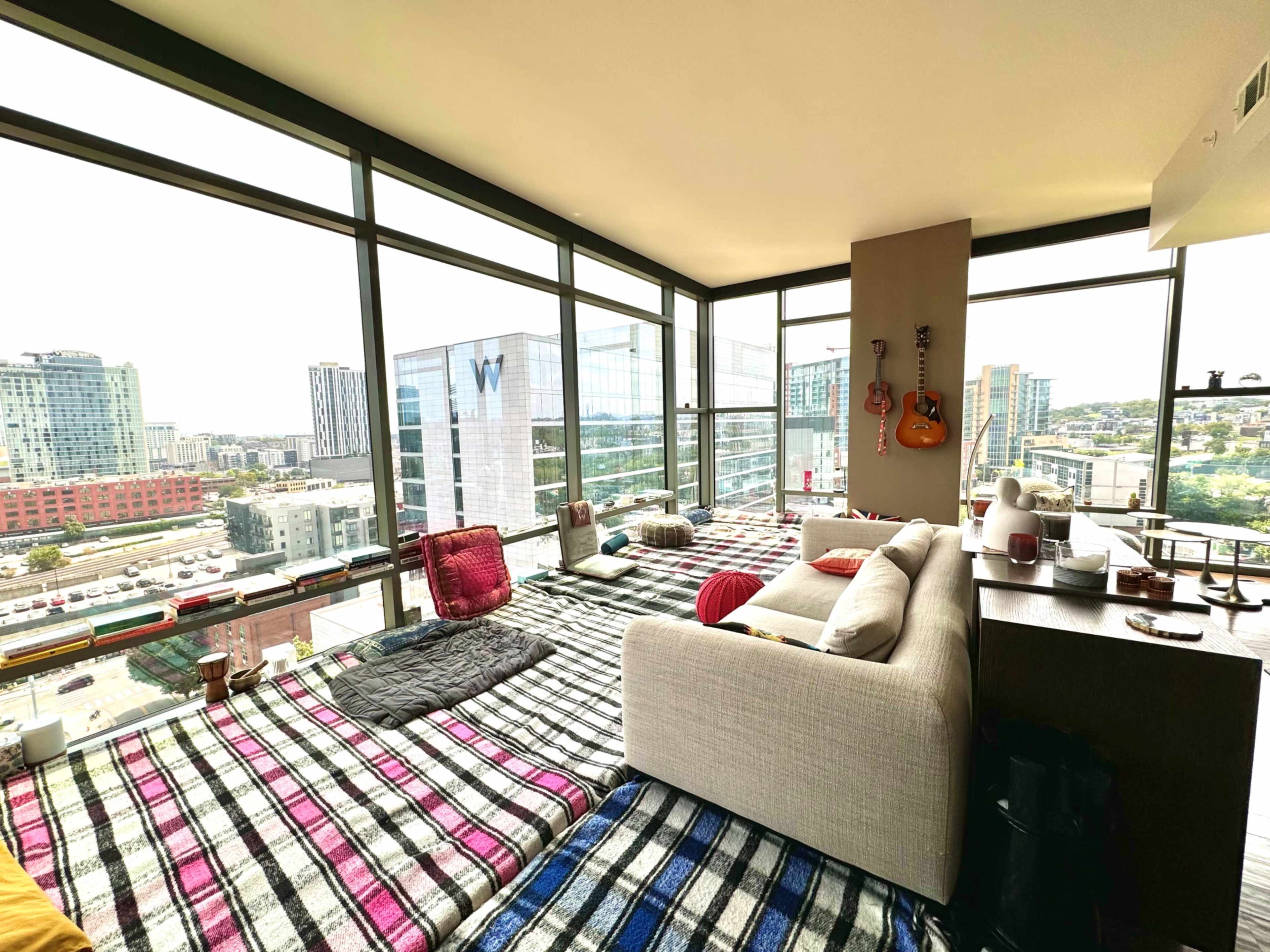 A modern living space features a large window with views of the city, furnished with plush blankets on the floor and a sectional sofa.