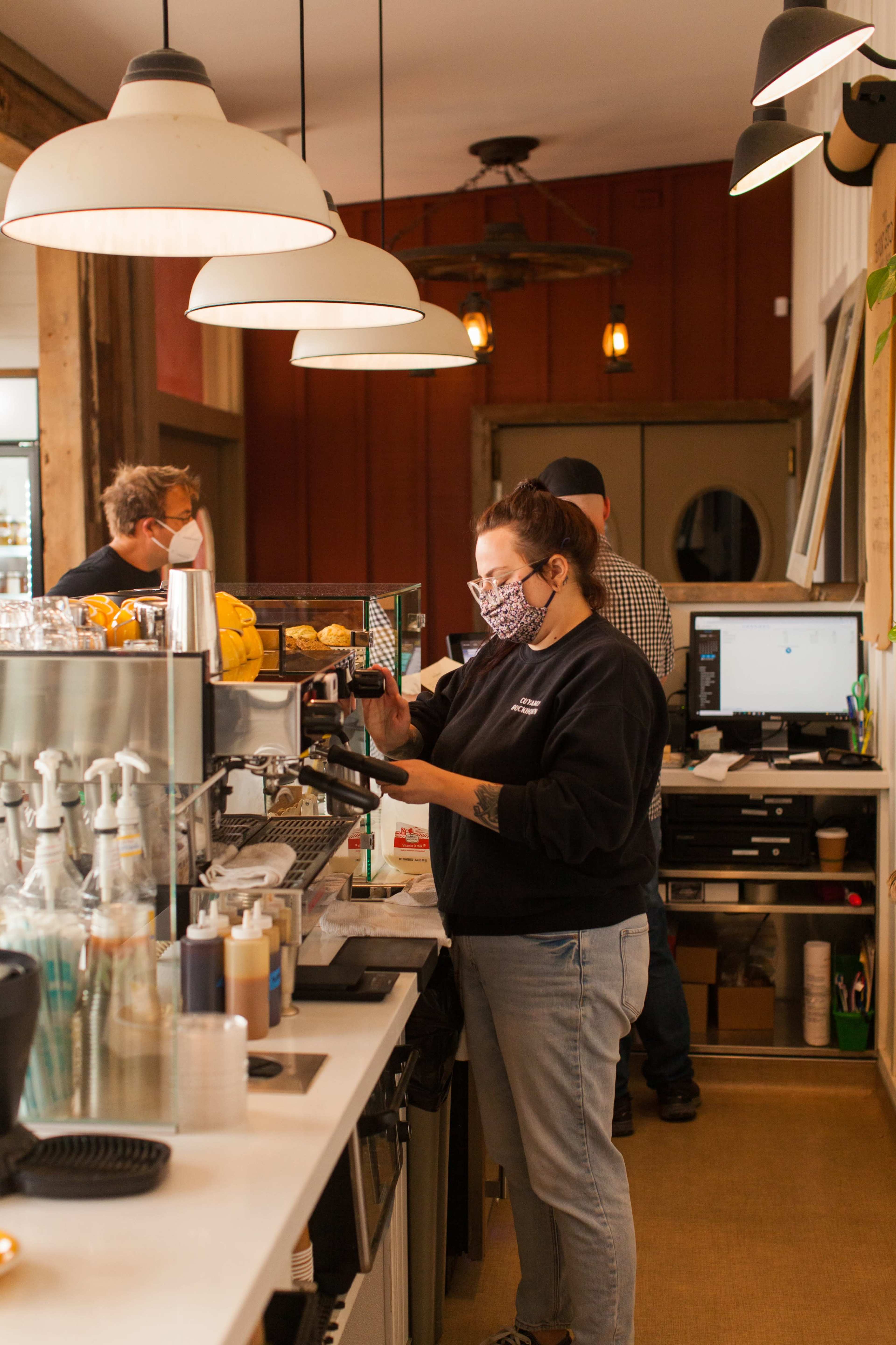 A barista prepares drinks at a coffee shop, while another employee organizes supplies in the background.
